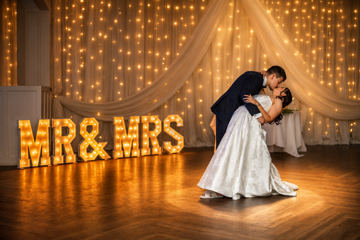 Bride and groom share first dance in front of illuminated MR & MRS marquee letters with fairy light backdrop.