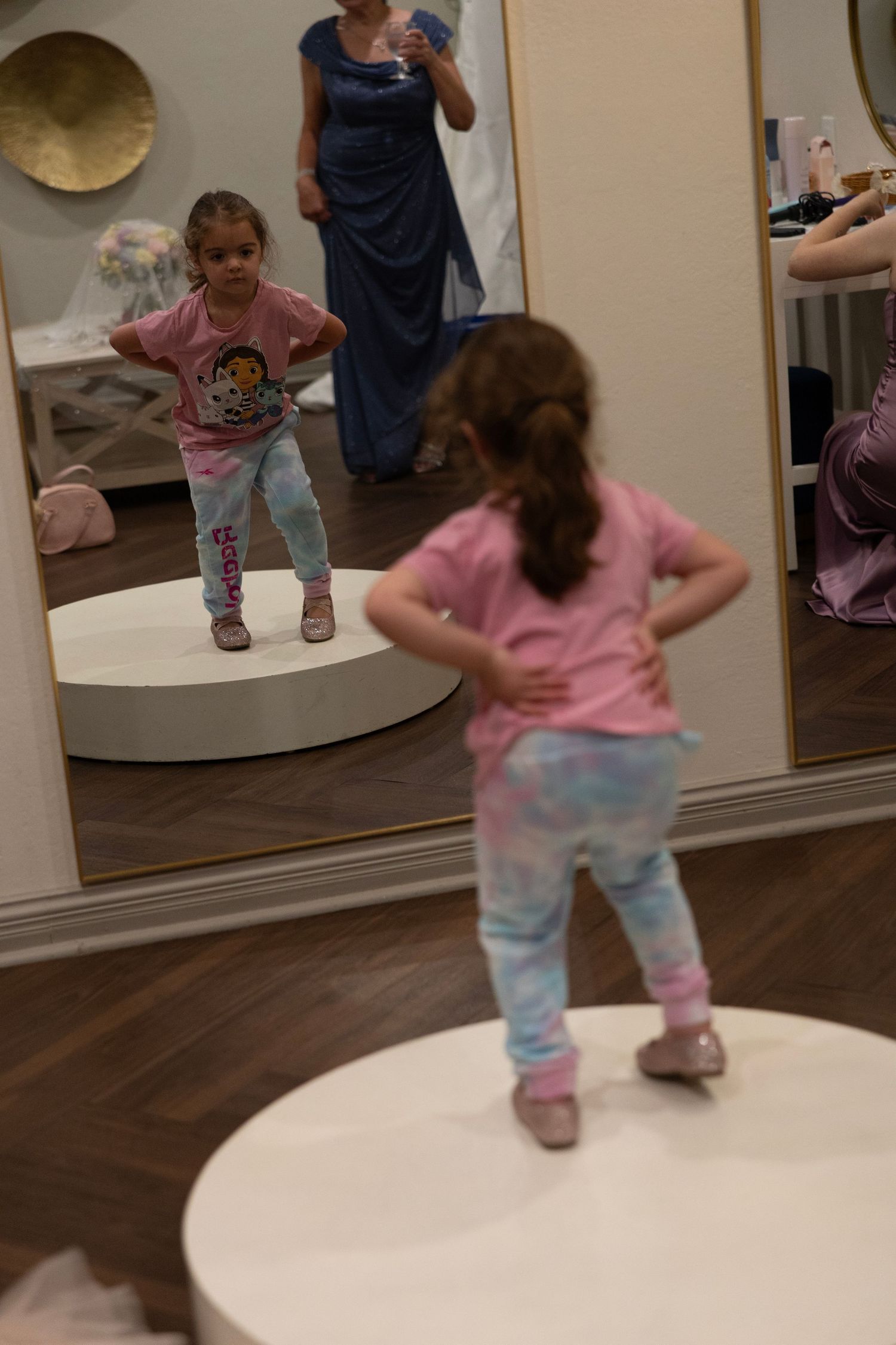Young girl in pink shirt and tie-dye pants poses confidently in front of a mirror in a clothing store.