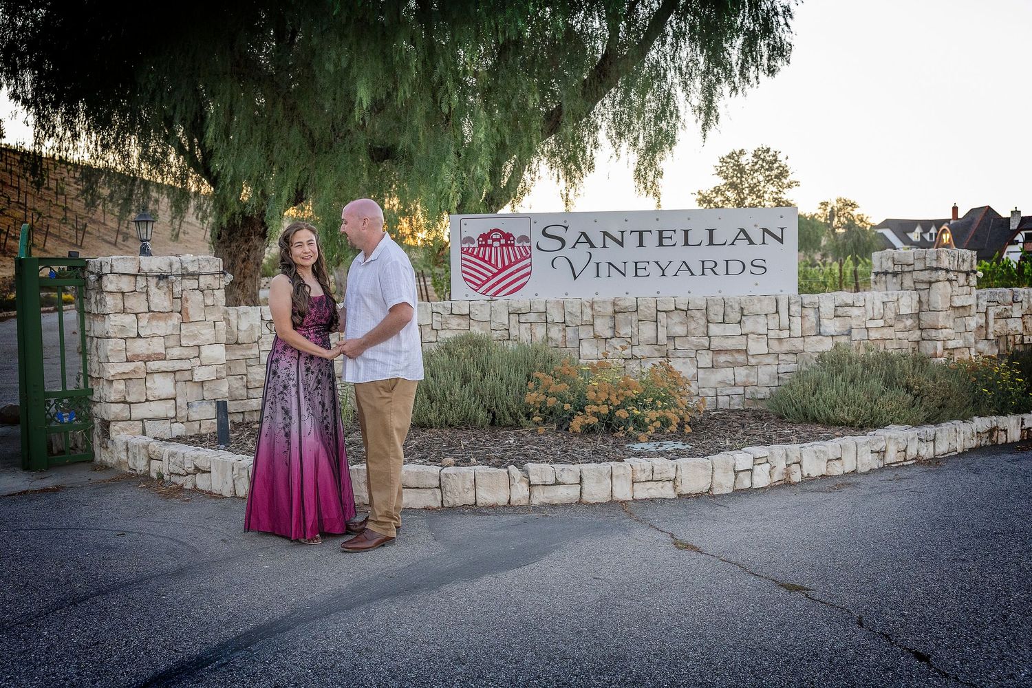 Couple posing in front of Santellan Vineyards sign surrounded by lush trees and stone wall at golden hour.