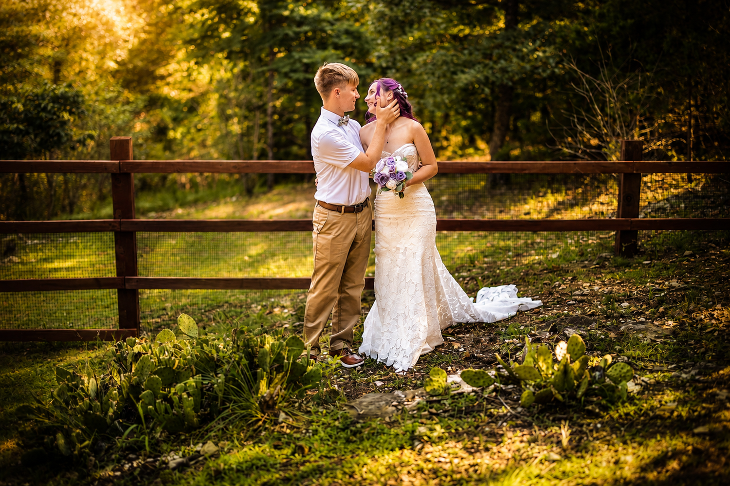 Bride and groom share a romantic moment by a rustic wooden fence surrounded by lush greenery in golden sunlight.
