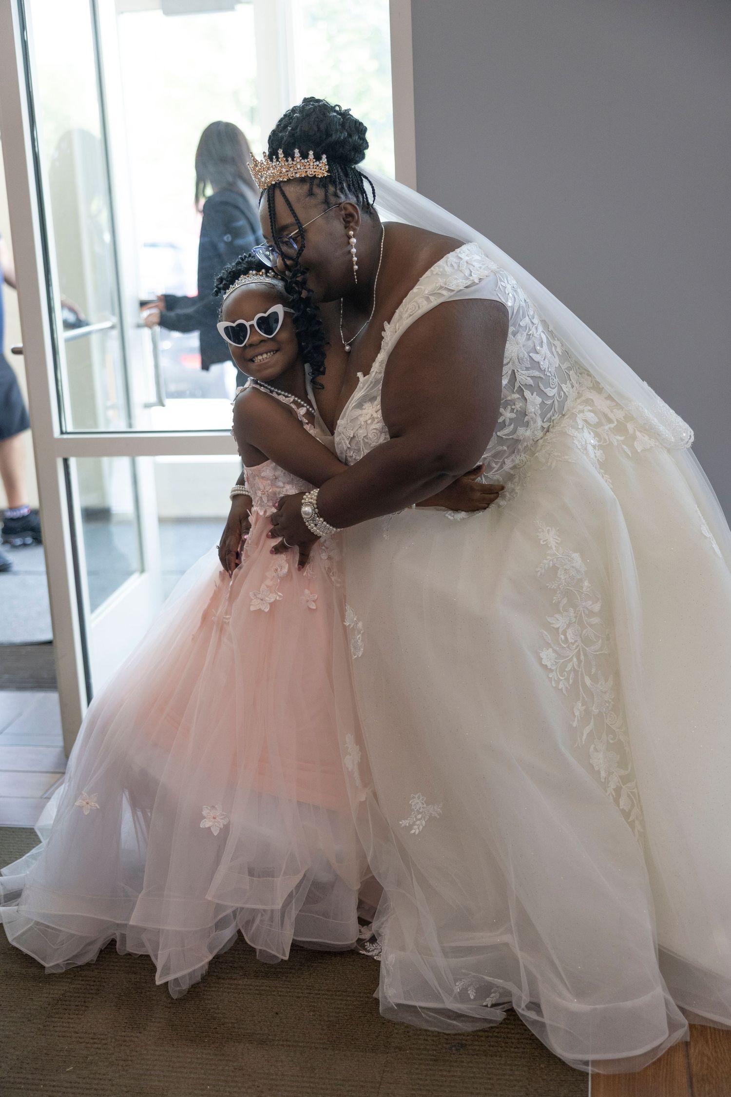 Bride in white lace gown hugs flower girl in pink tulle dress, both wearing crowns at a wedding venue.