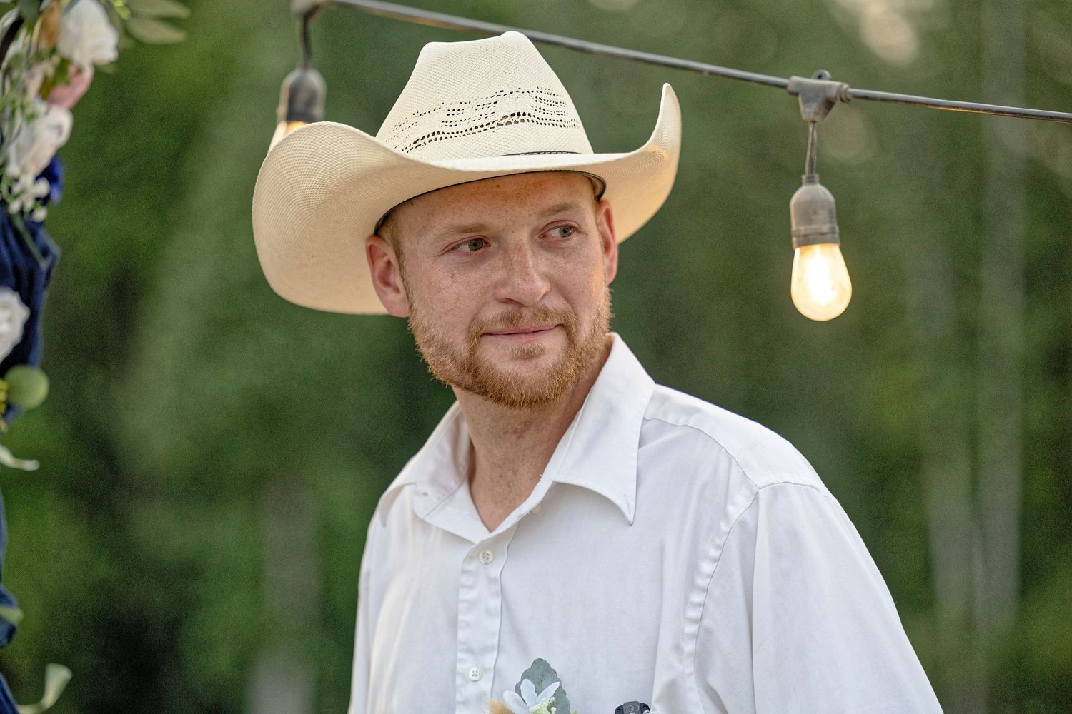Man wearing a straw cowboy hat and white shirt at an outdoor event with string lights and floral decor.