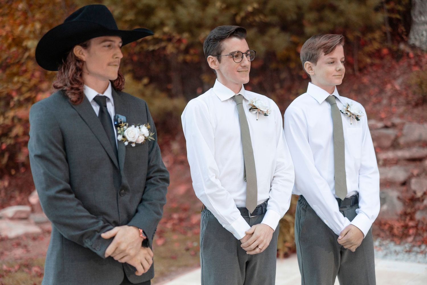 Three groomsmen stand in formal attire with boutonnieres against a backdrop of autumn foliage.