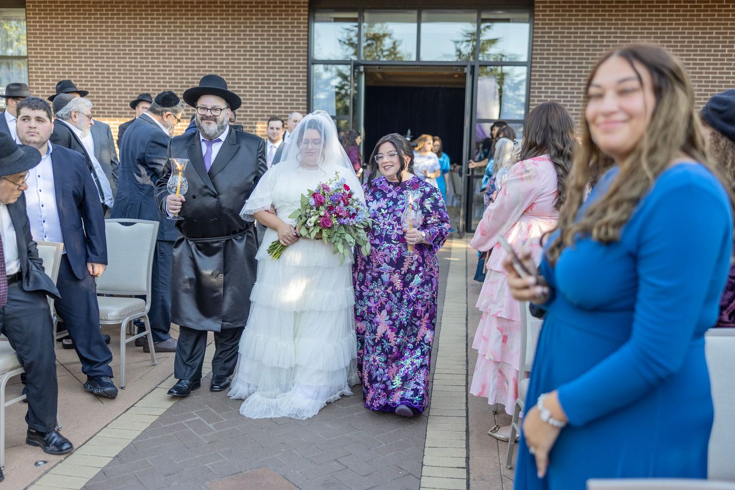 Wedding guests gather outside a brick building during a celebration, sharing candid moments and conversations.