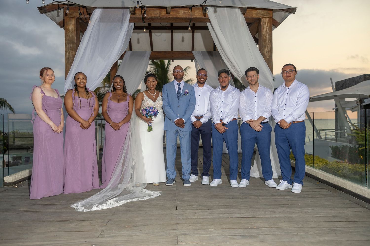 Wedding party poses together under wooden arch at outdoor venue with bridesmaids in pink dresses and groomsmen in blue.