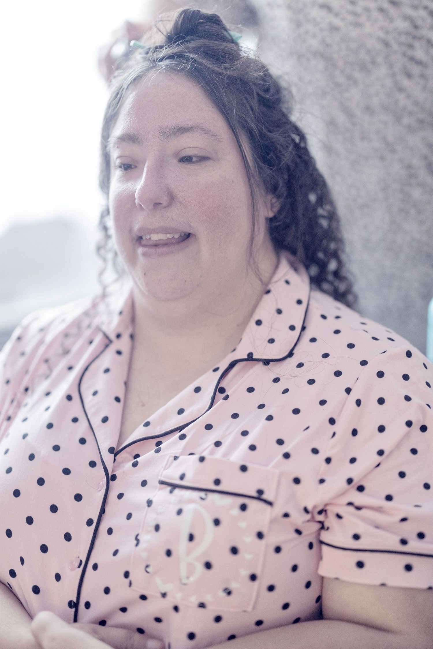 A person wearing a pink polka dot pajama top smiles warmly while relaxing in bed.