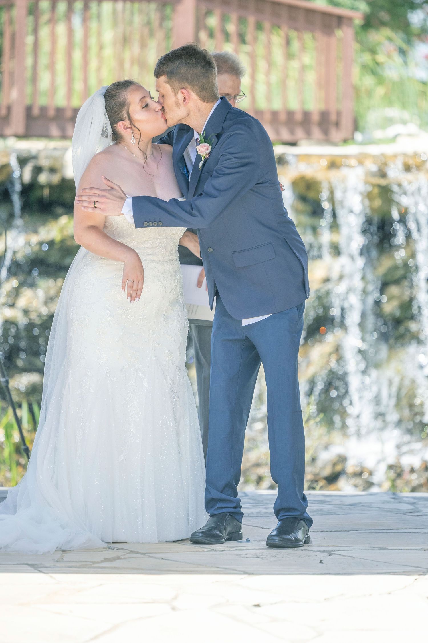 A bride in a white gown and groom in navy suit share a romantic kiss under a rustic wooden pavilion.