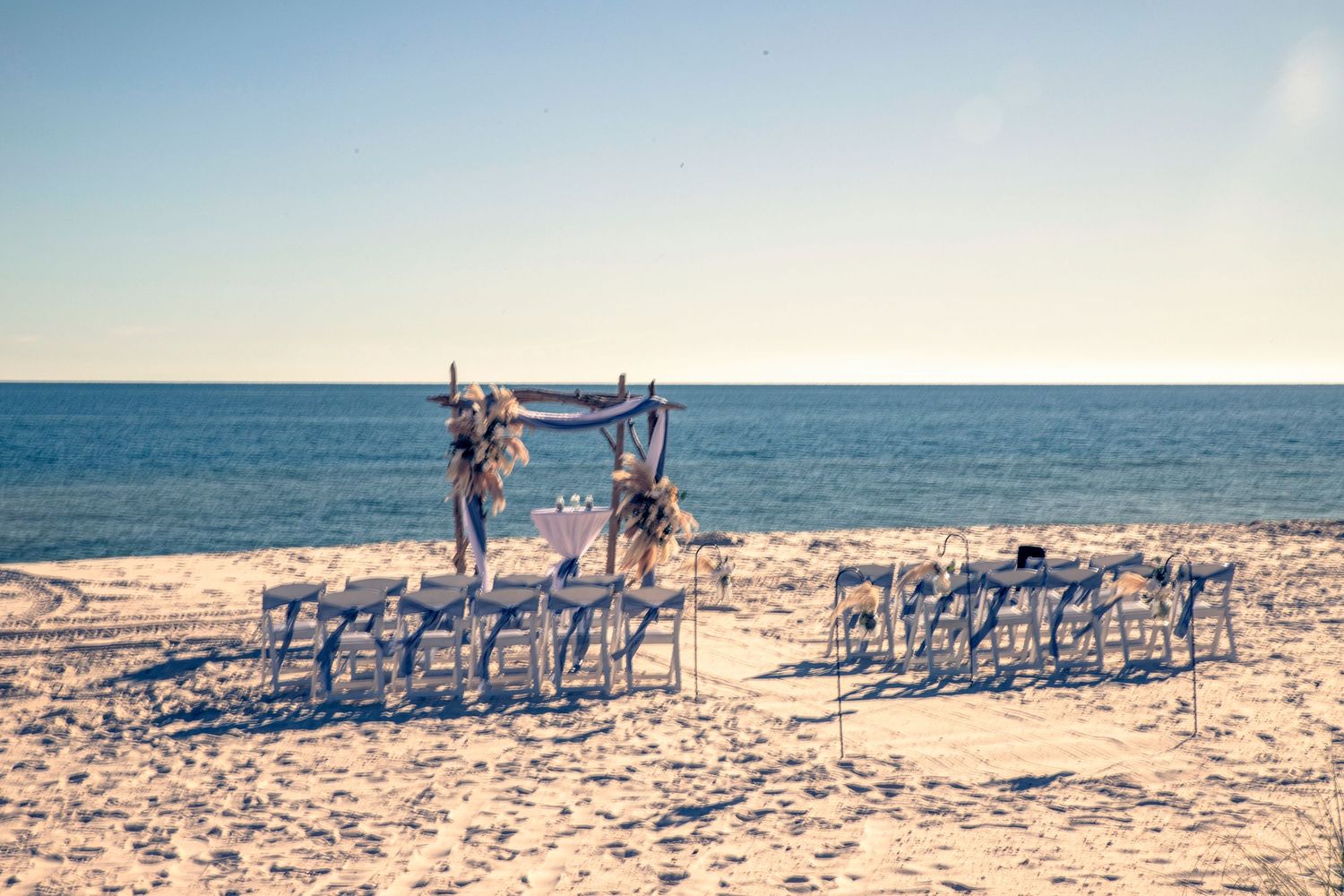 White chairs arranged on a sandy beach for a seaside wedding ceremony with ocean views at sunset.