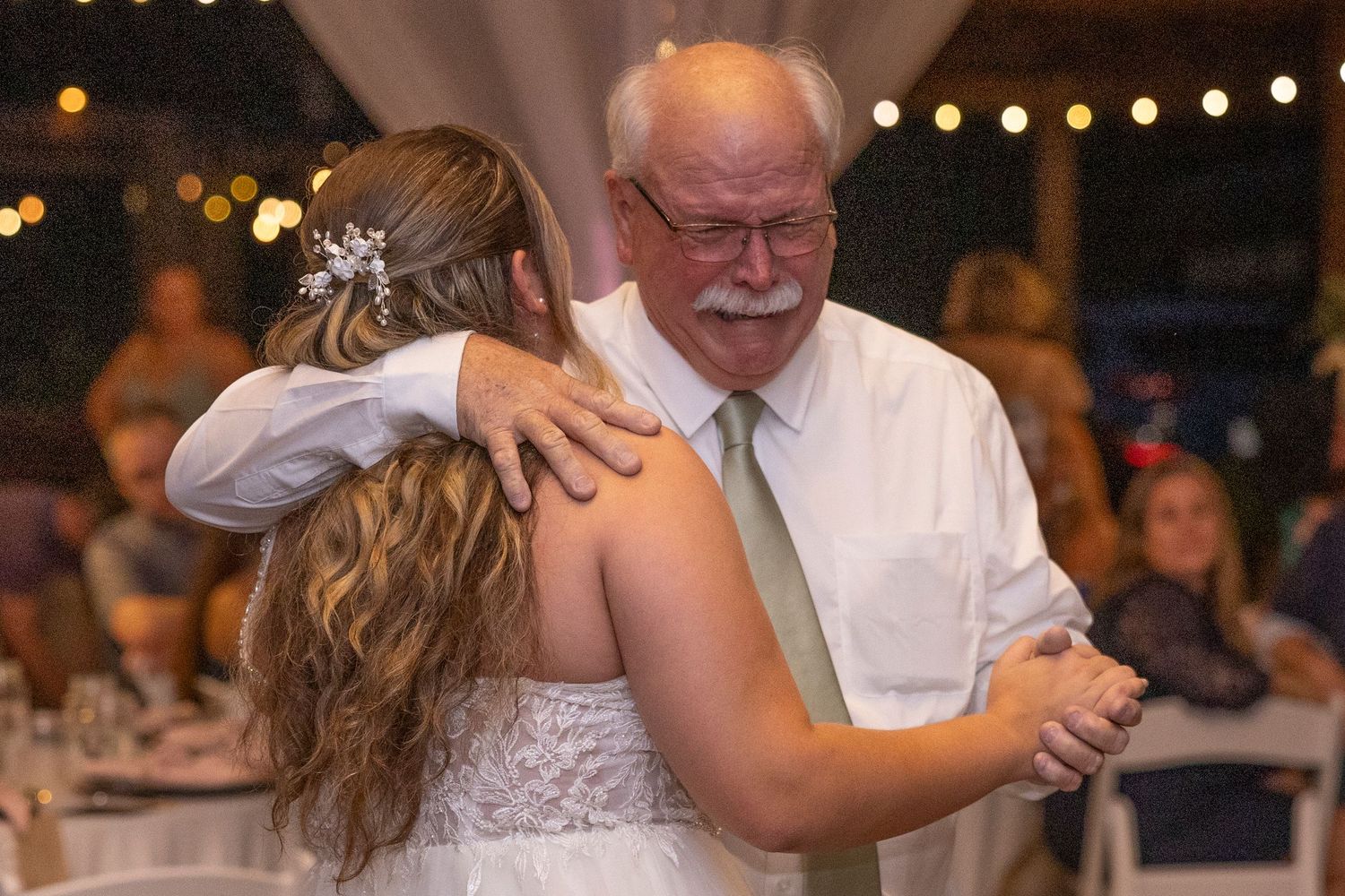 A tender moment captured during a father-daughter dance at a wedding reception with string lights in the background.