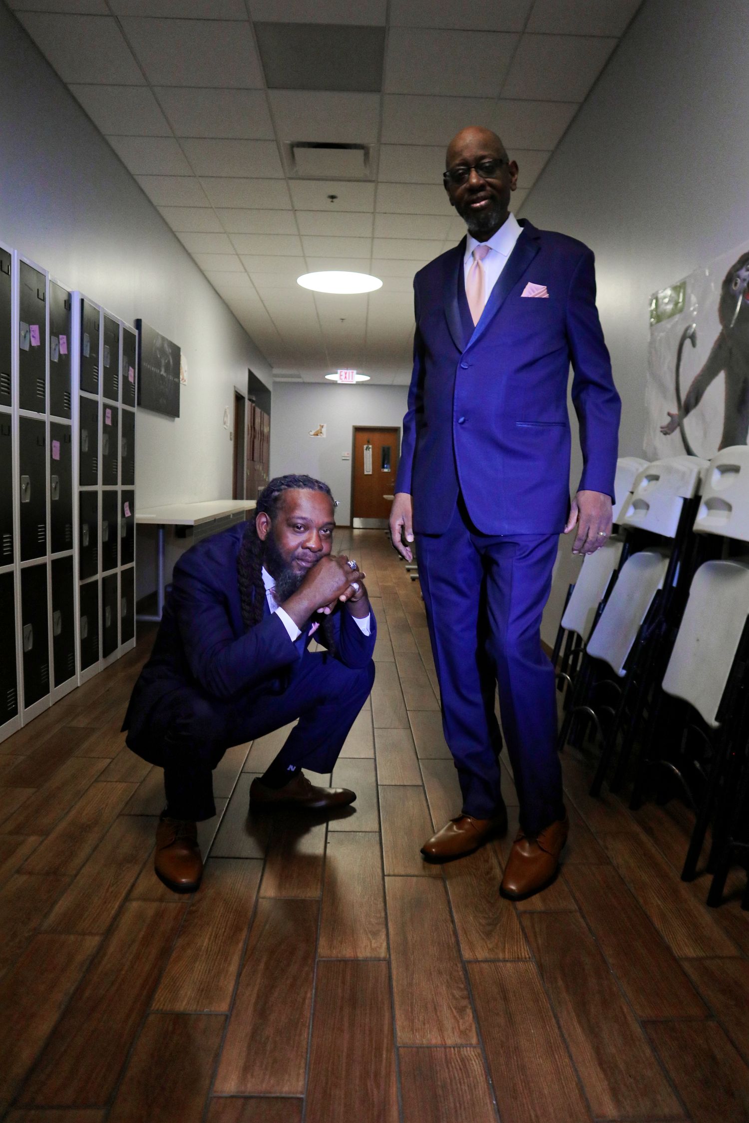 Two well-dressed men in matching blue suits pose in a hallway, one crouching and one standing tall.