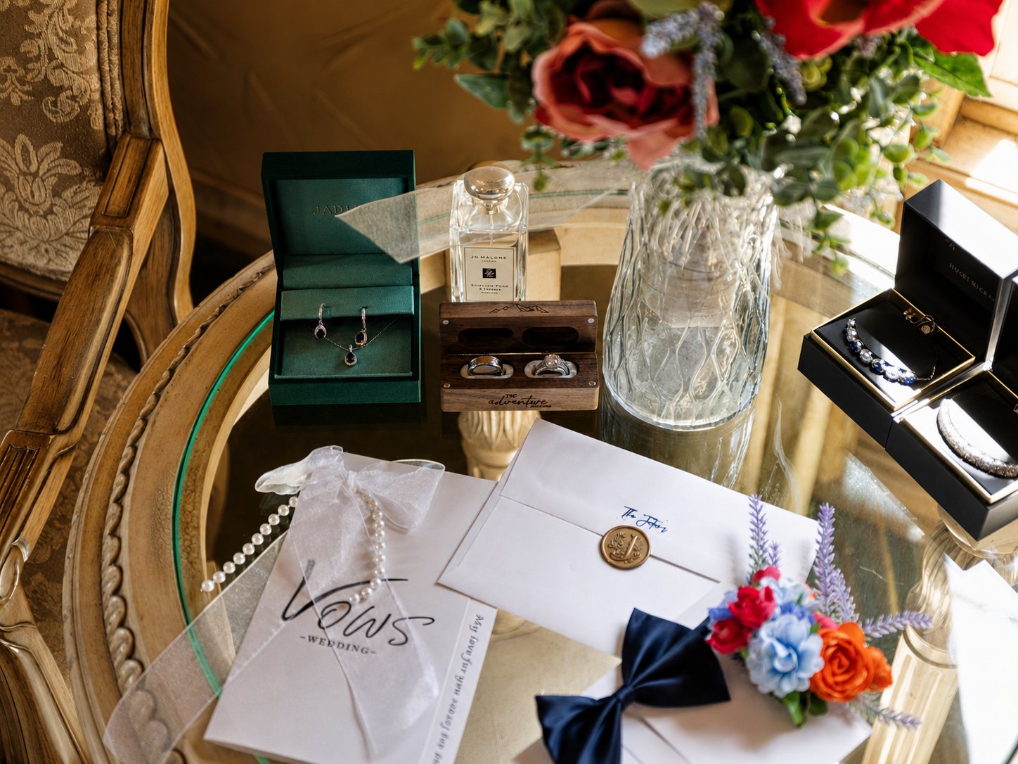 Wedding flat lay with jewelry boxes, rings, bow tie, floral bouquet, and Vows envelope on gold table.