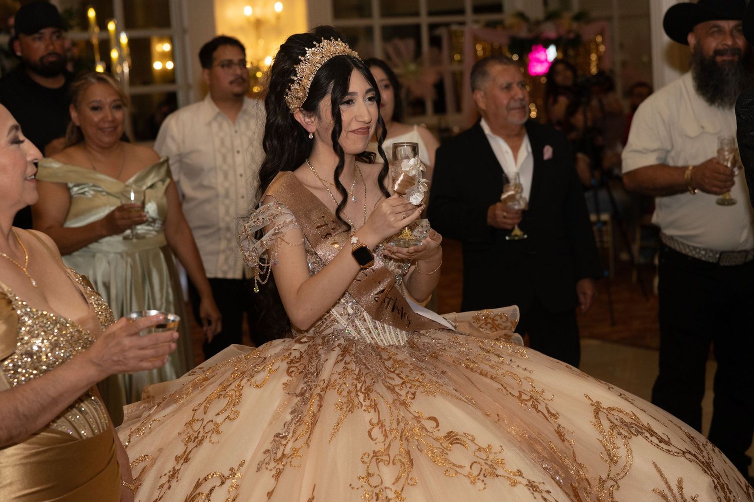 Young woman in gold quinceañera ball gown with tiara holds a gift at her elegant celebration party.