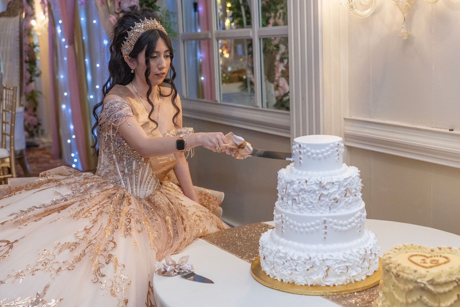 Young woman in ornate gold gown cutting a white tiered wedding cake at an elegant celebration.