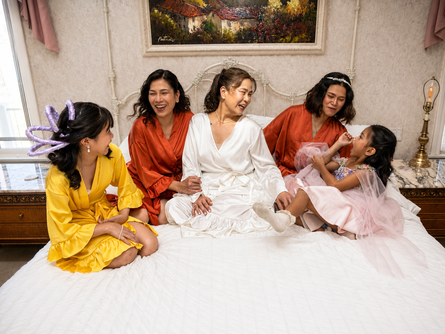 Bride in white robe laughing with bridesmaids and flower girls in colorful robes on a bed.
