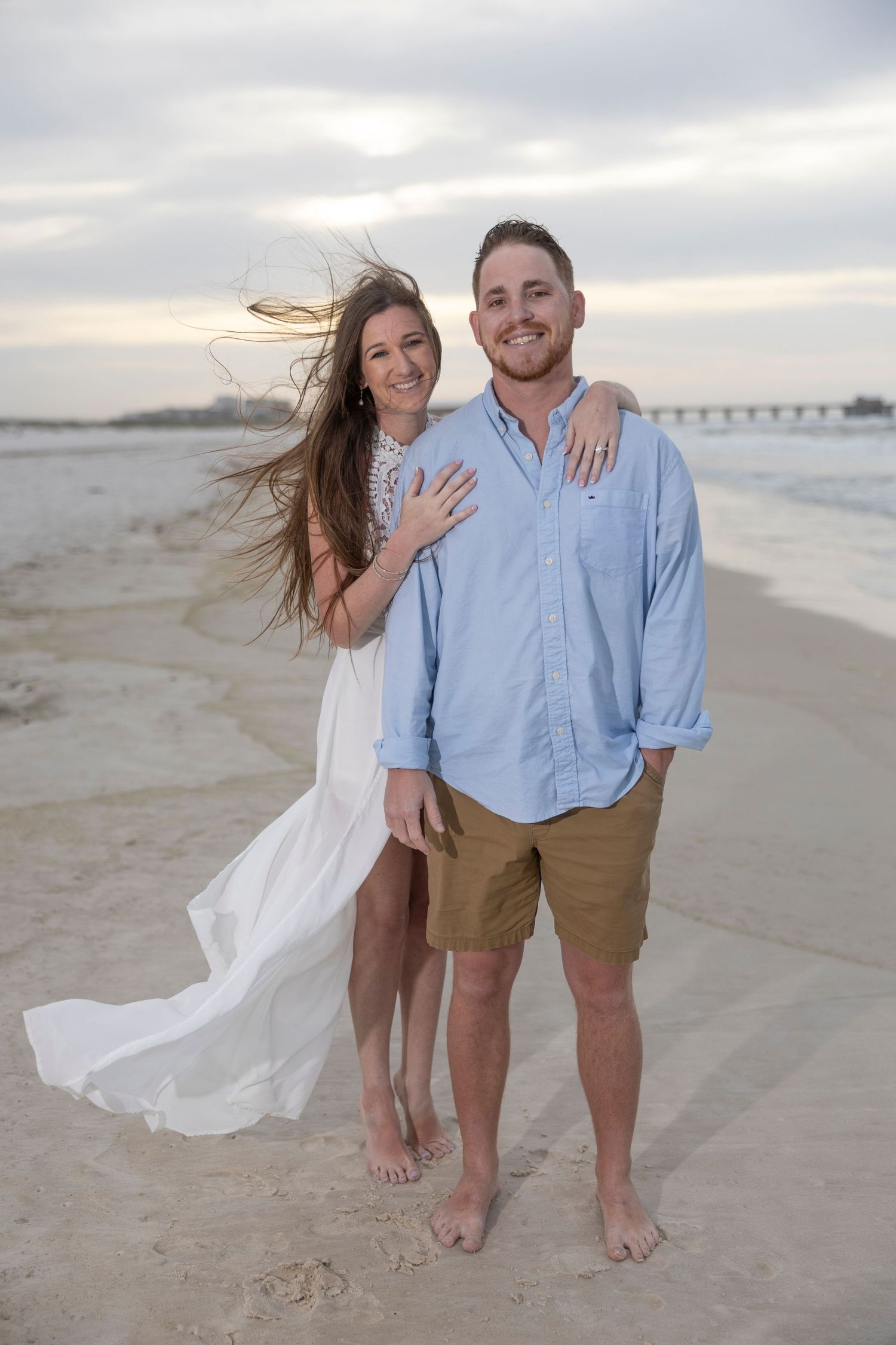 Happy couple posing on a sandy beach at sunset, woman in white dress with windswept hair, man in blue shirt.
