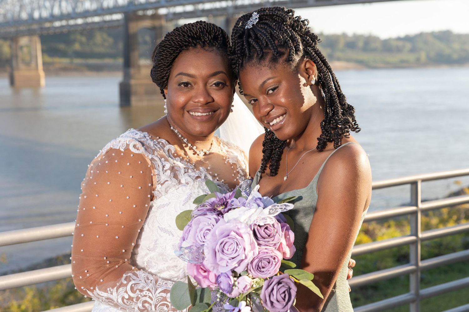 Two women in elegant attire pose with purple roses bouquet near a river bridge at a wedding celebration.