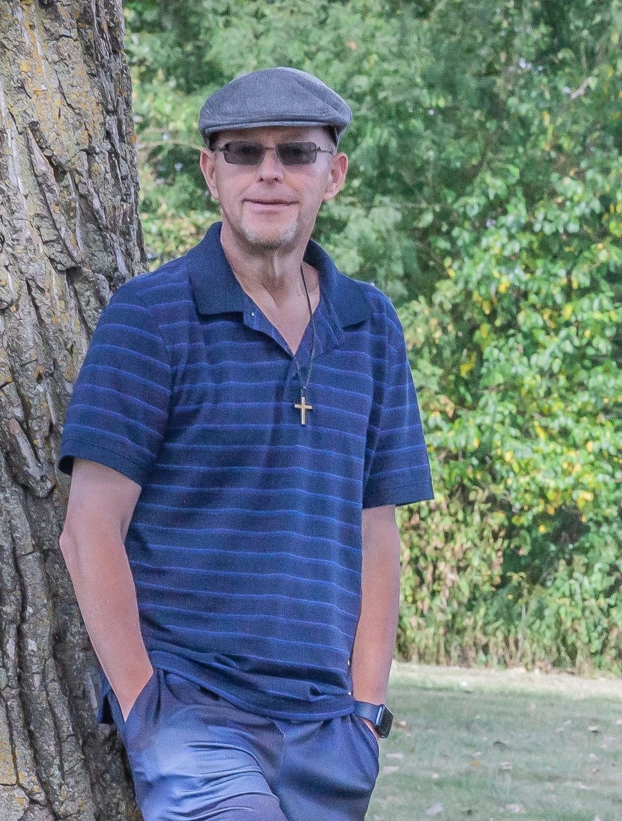 Man in flat cap and sunglasses wearing a cross necklace, leaning against a tree in a lush green outdoor setting.