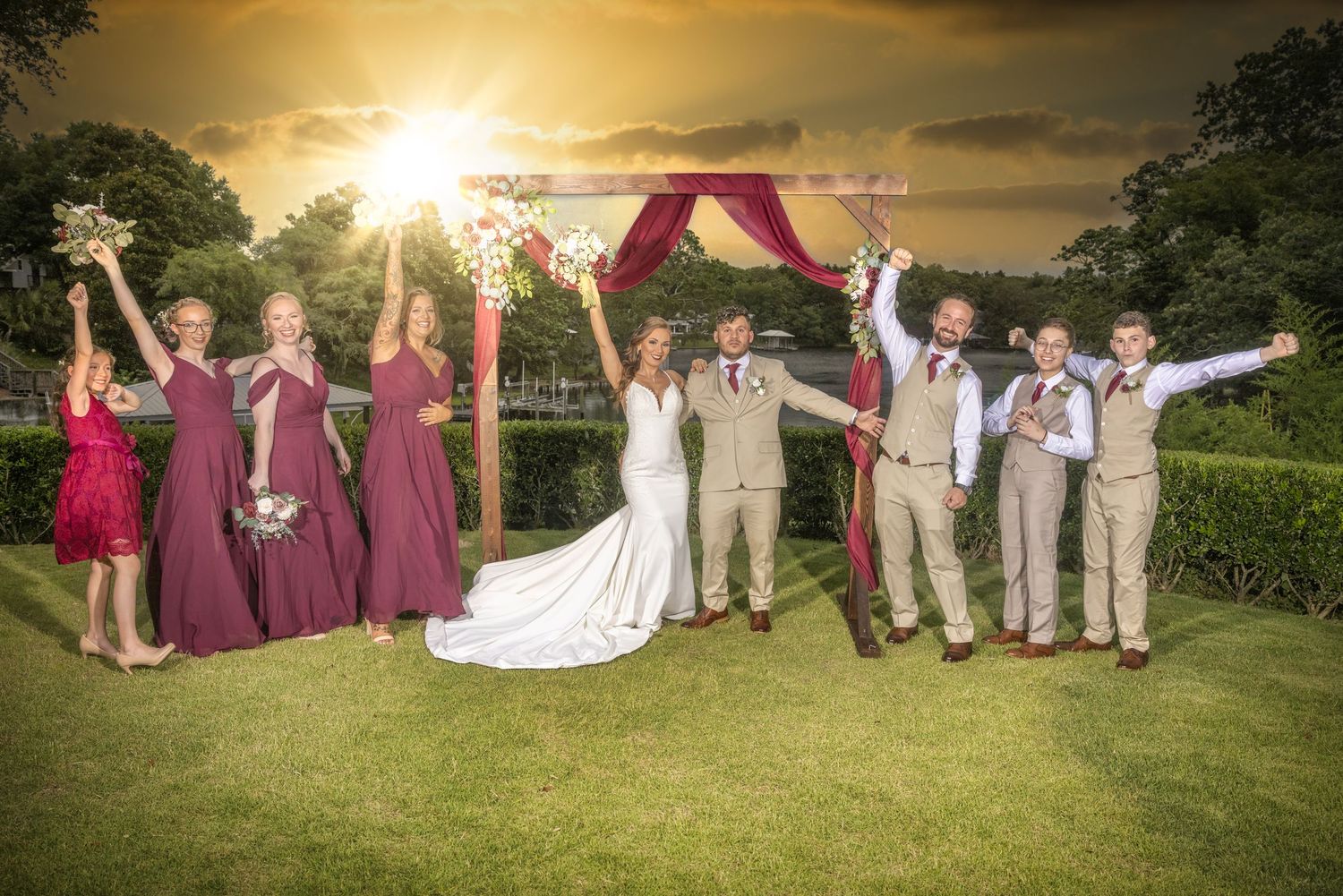 Wedding party celebrates under floral arch during golden hour with bridesmaids in burgundy dresses.