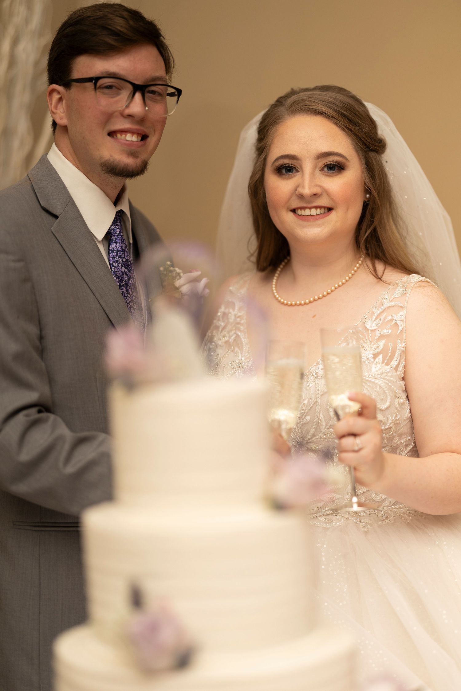 Wedding couple stands beside elegant white tiered cake at reception.
