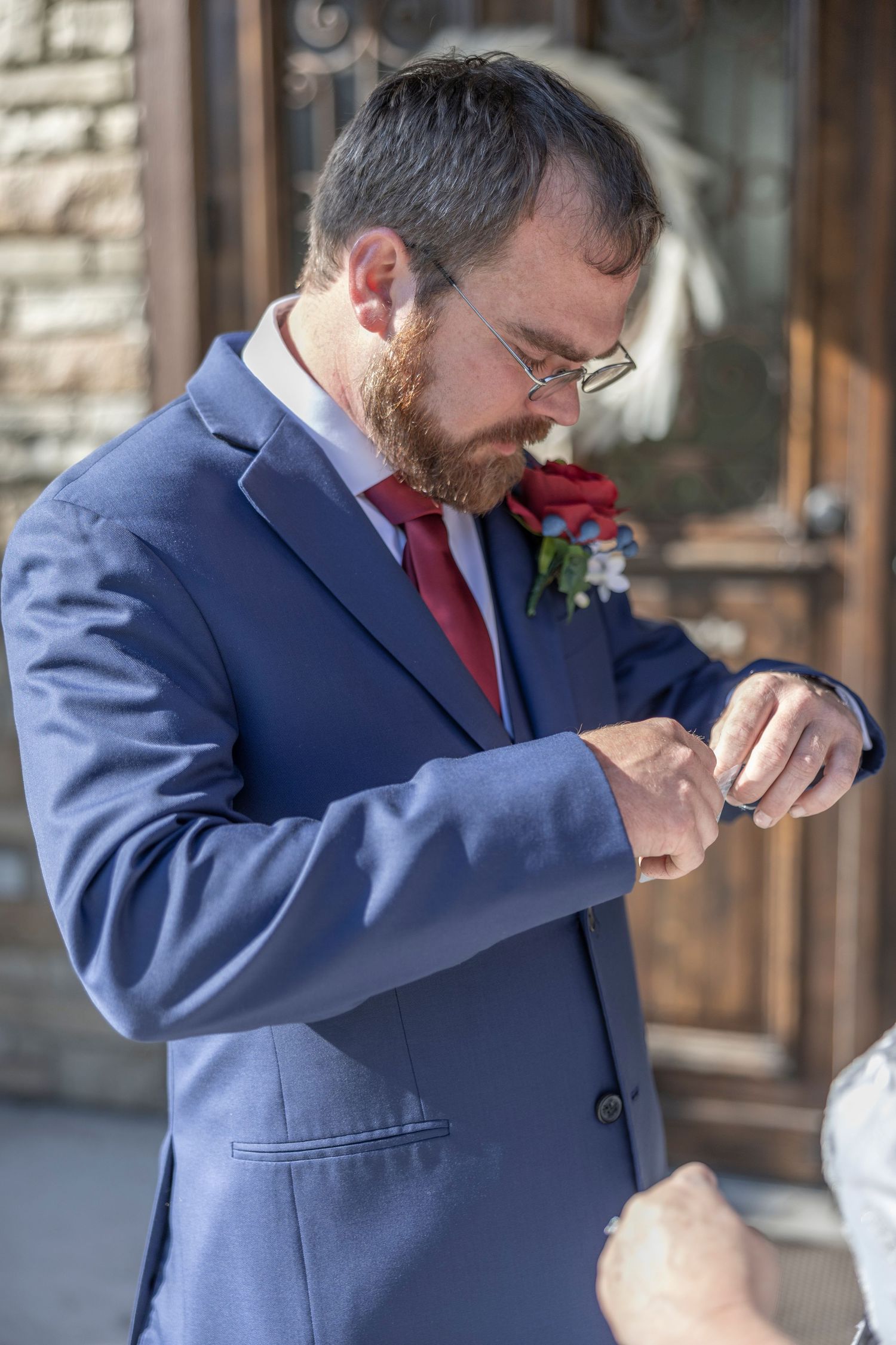 A person in a navy suit and red tie smells a boutonniere flower on their lapel.