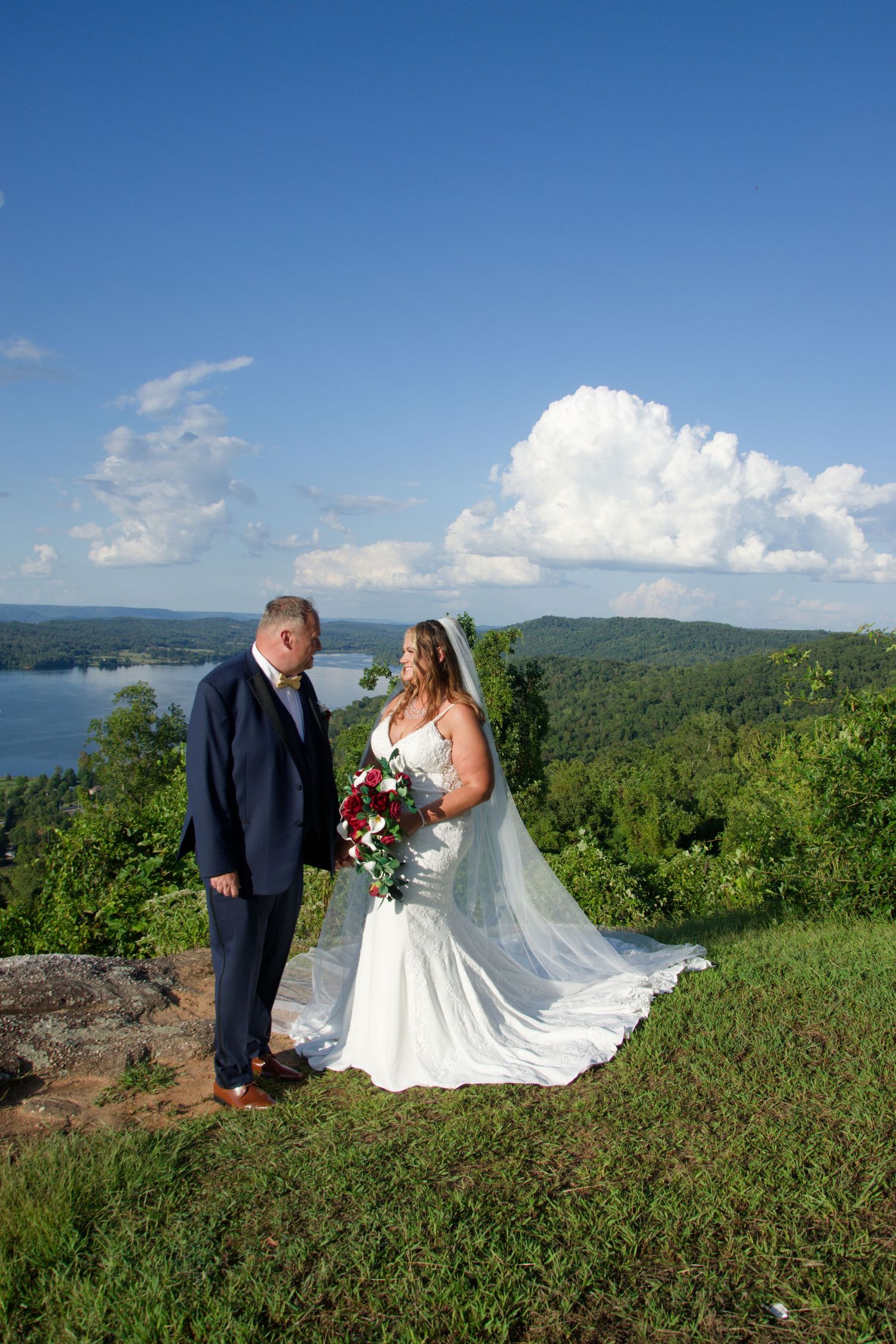 Couple in wedding attire pose together on a scenic mountaintop overlook with rolling green hills and blue skies in background.