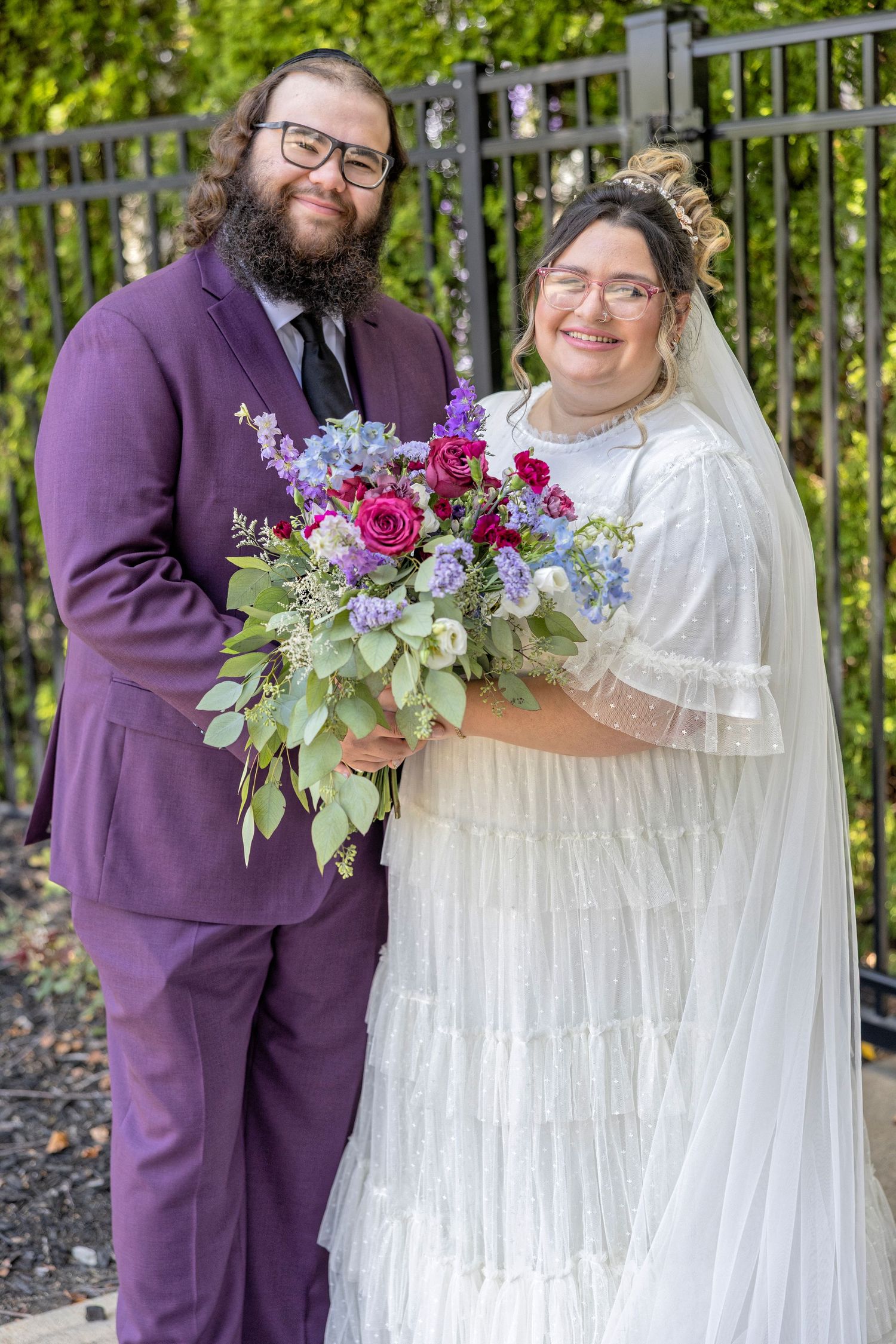 Wedding couple poses together outdoors in purple and white attire with vibrant floral bouquet against garden backdrop.