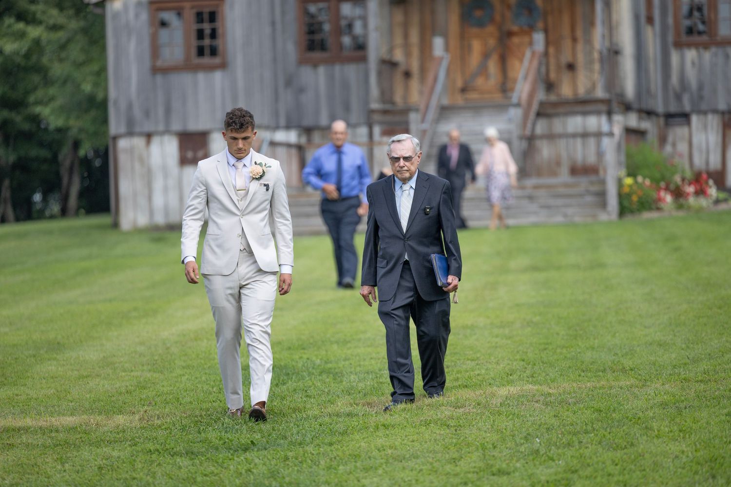 A group of people walk across a lush green lawn near a rustic wooden building on a sunny day.