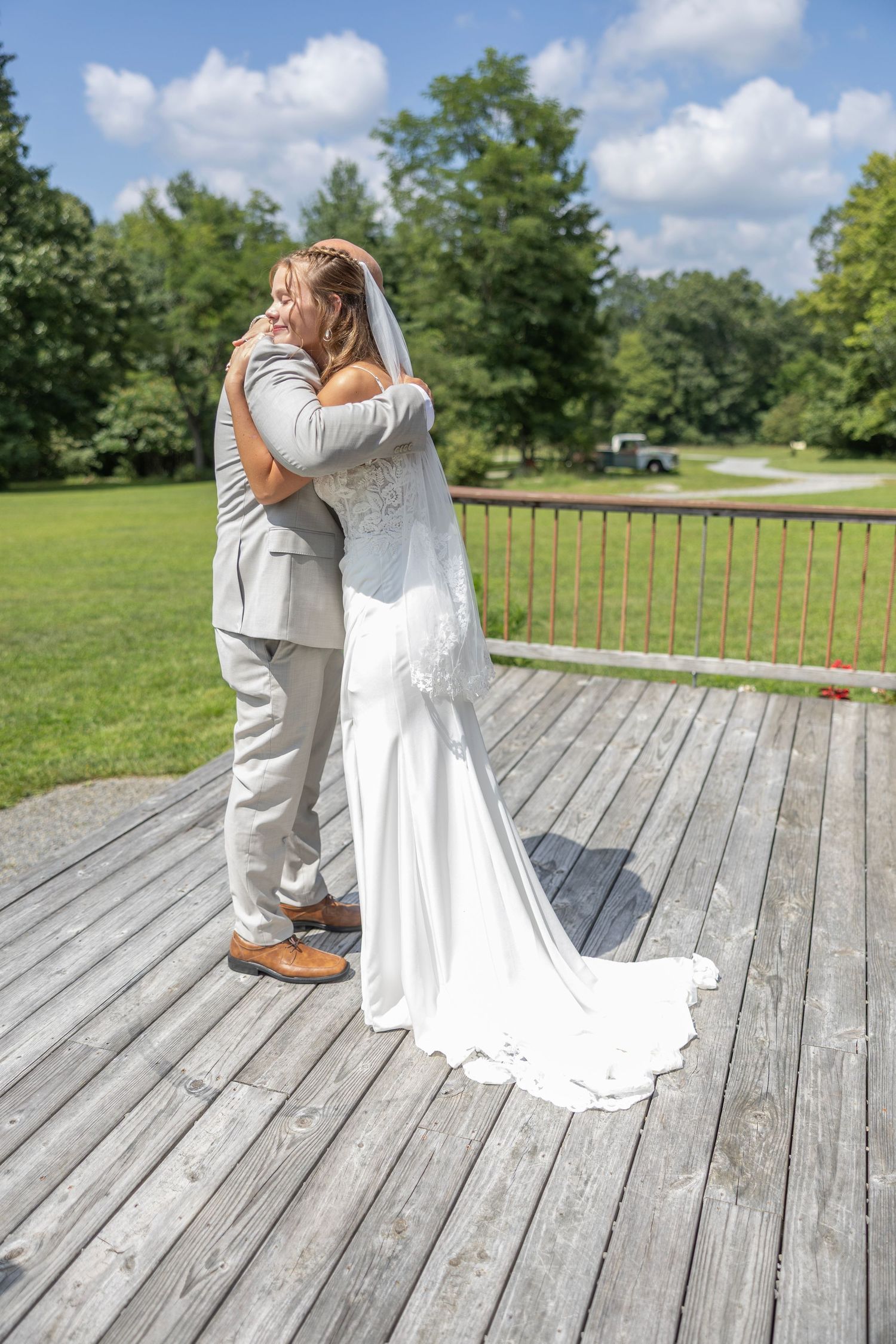 A couple shares an intimate embrace on a wooden deck surrounded by green trees on their wedding day.