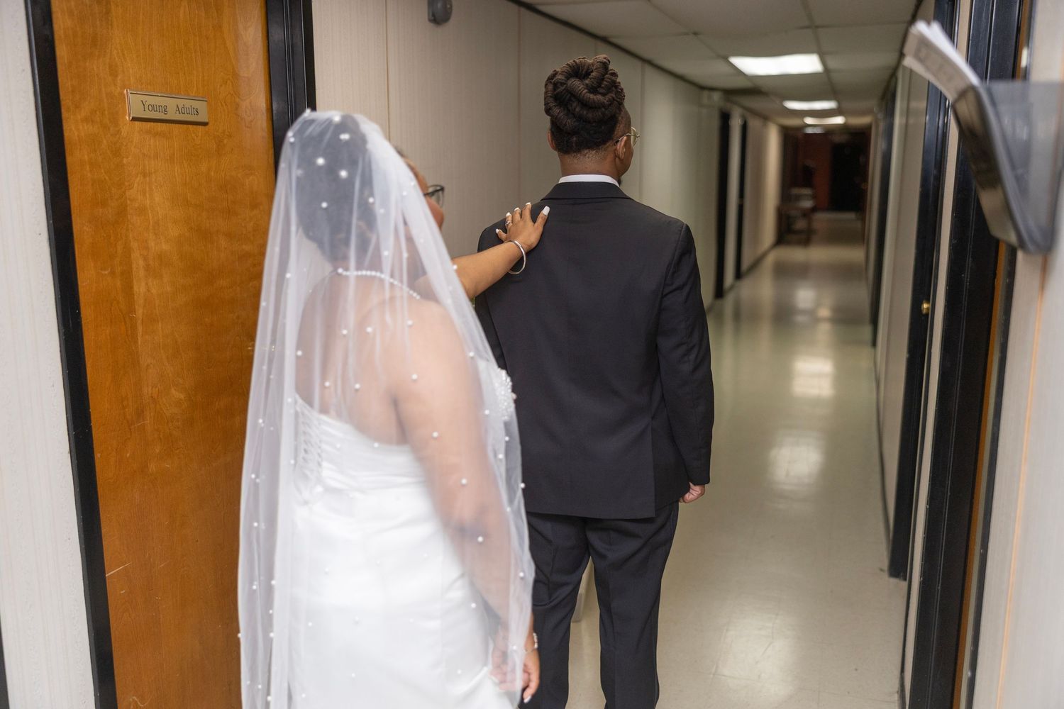A bride in a white veil and groom in a dark suit walk down a brightly lit hallway corridor.