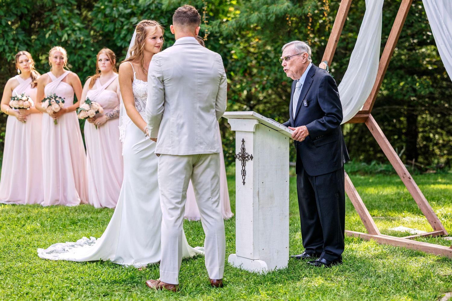 Wedding ceremony guests seated in white chairs on a lawn with pink bridesmaids dresses visible in the background.