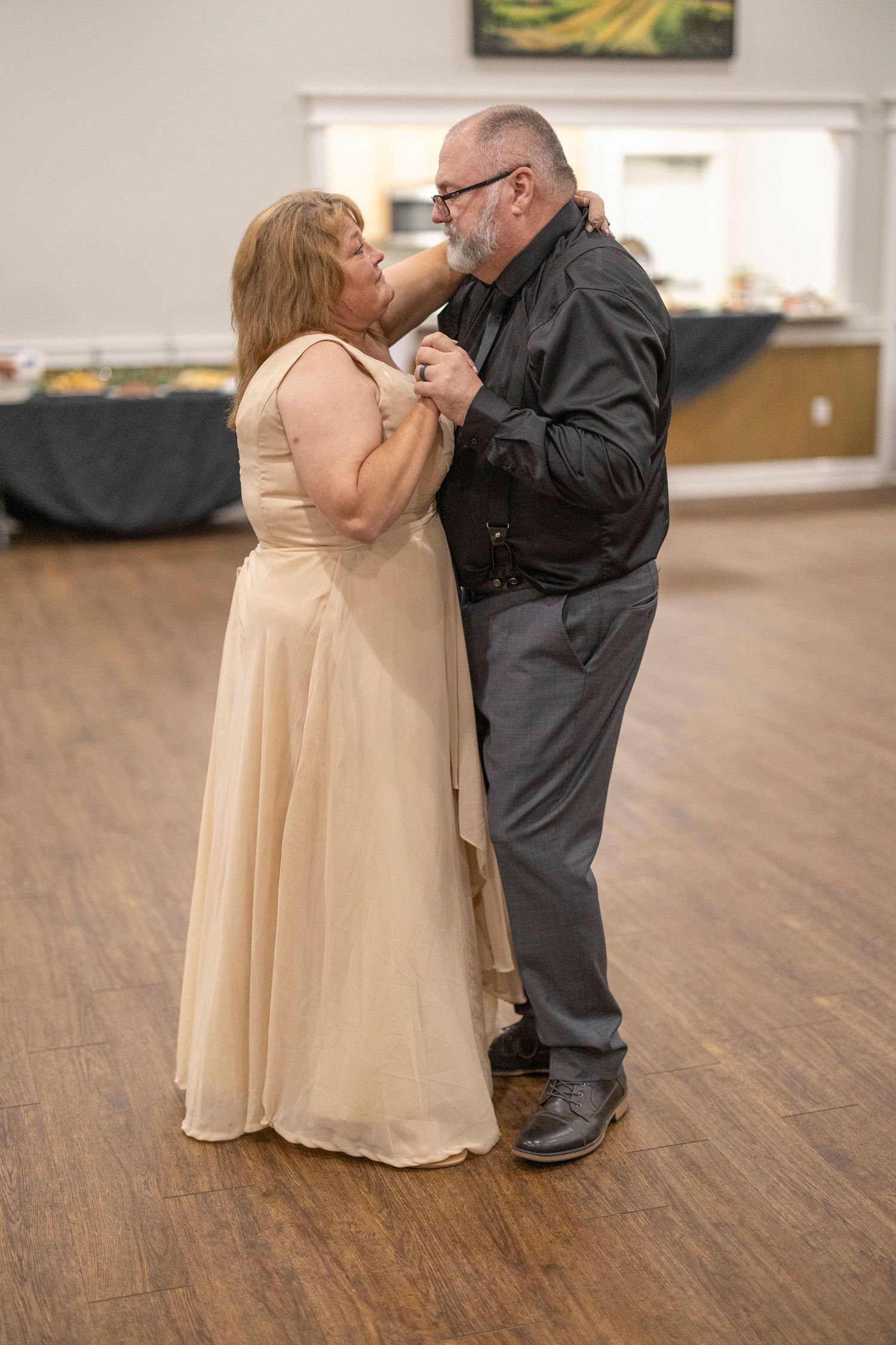 A couple shares a tender moment while dancing together on a hardwood floor, with the person in a flowing beige dress.