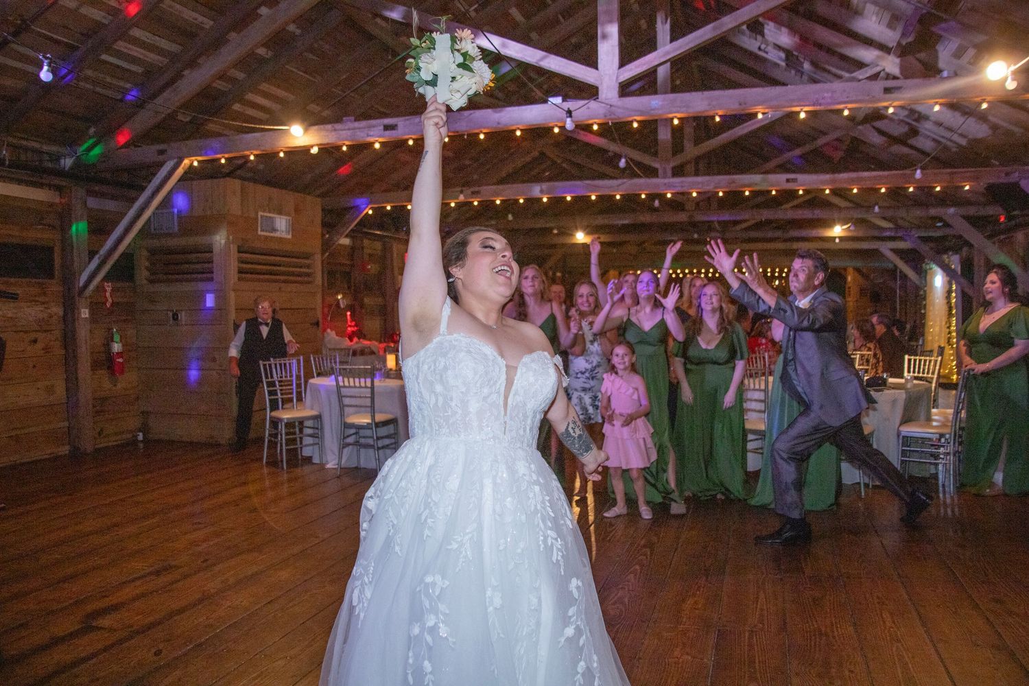 Wedding reception guests dance and celebrate under string lights in a rustic wooden barn venue.