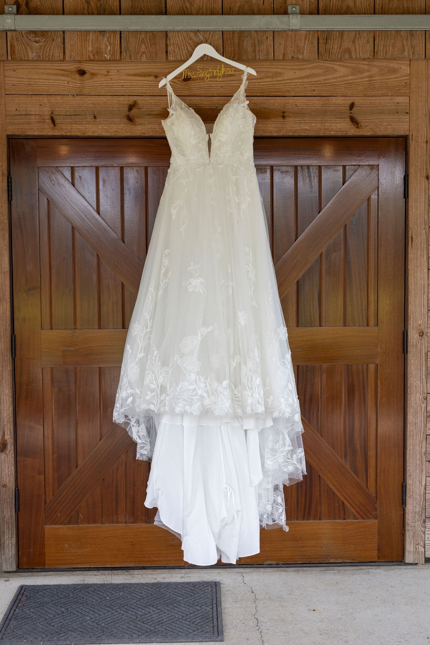 A white wedding dress with lace details hangs against a rustic wooden barn door backdrop.