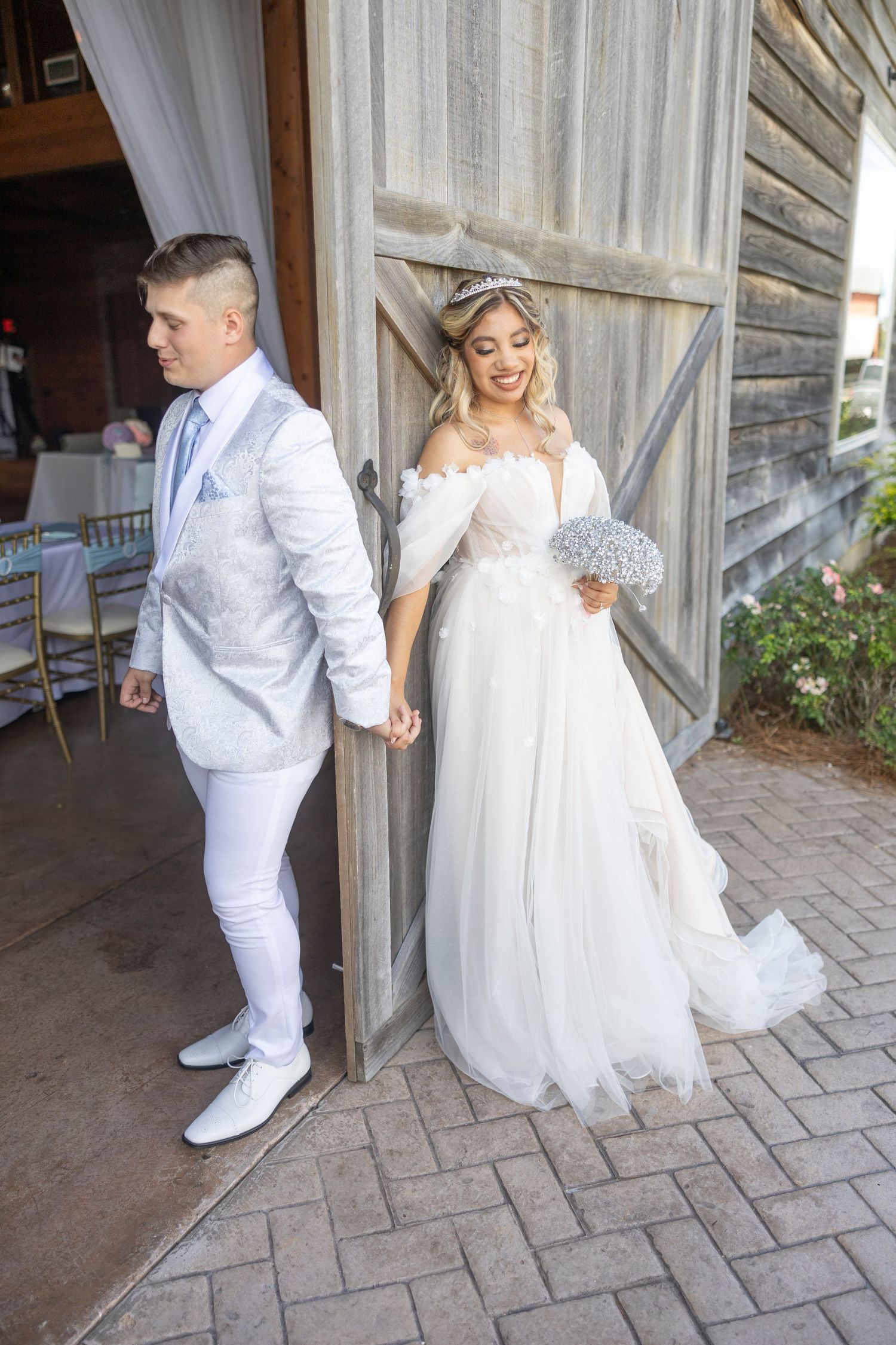 A couple in all-white wedding attire hold hands while standing by a rustic barn door on their special day.