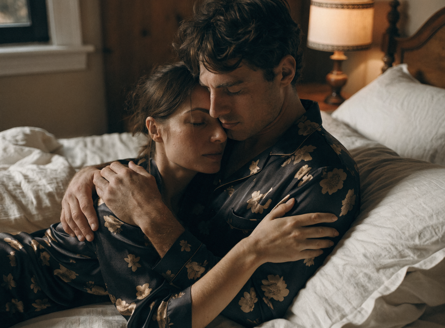 Couple in floral silk pajamas embracing tenderly on a cozy bed with white pillows and warm lamp light.