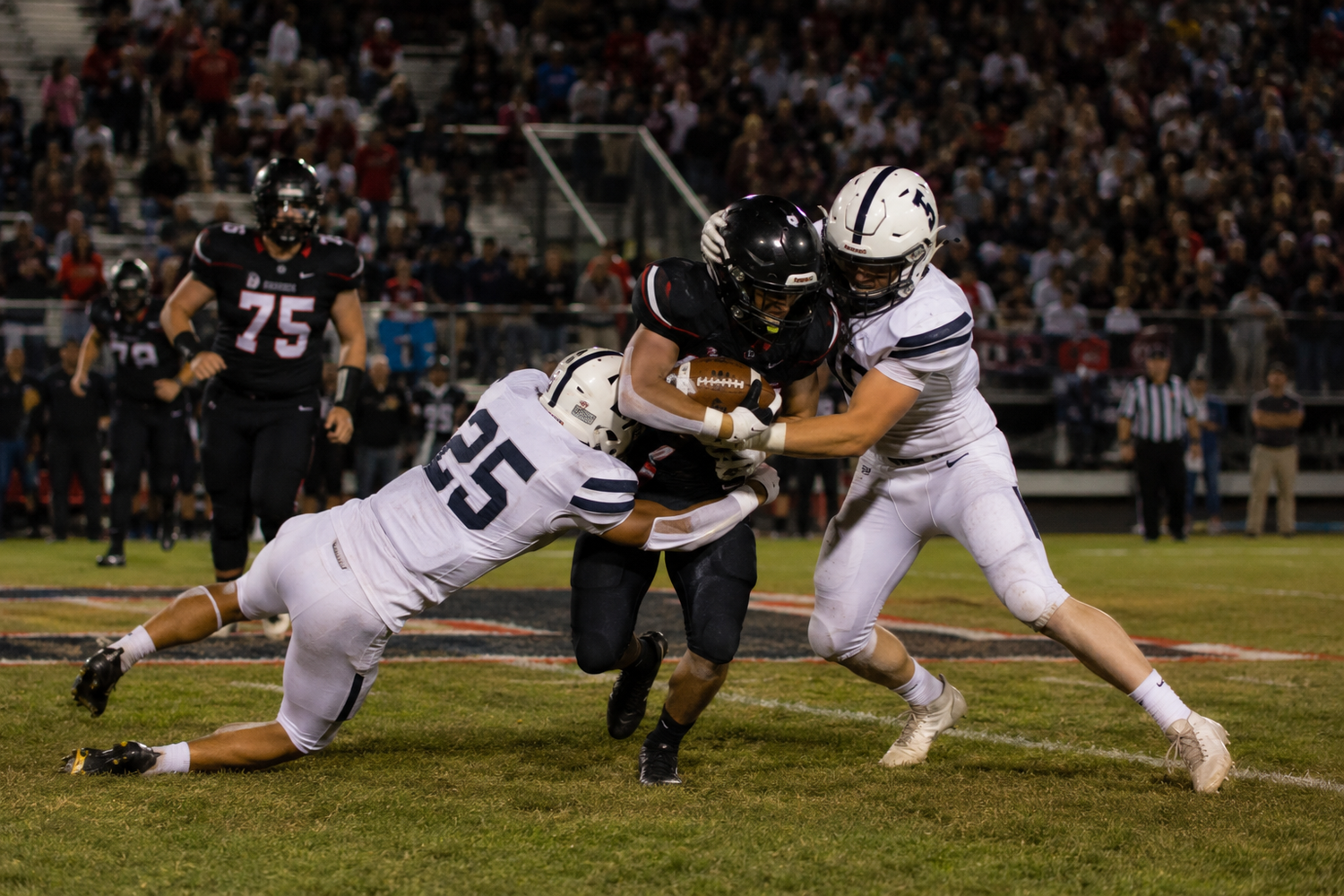 Two football players in white uniforms tackle an opposing player during an intense high school football game at night.