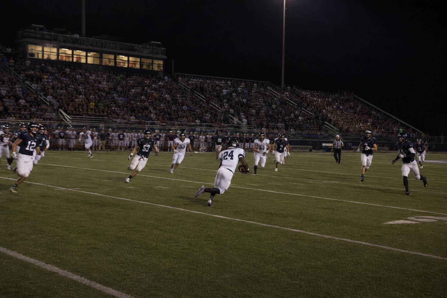 Football players compete during a night game on a well-lit stadium field with fans in the stands.