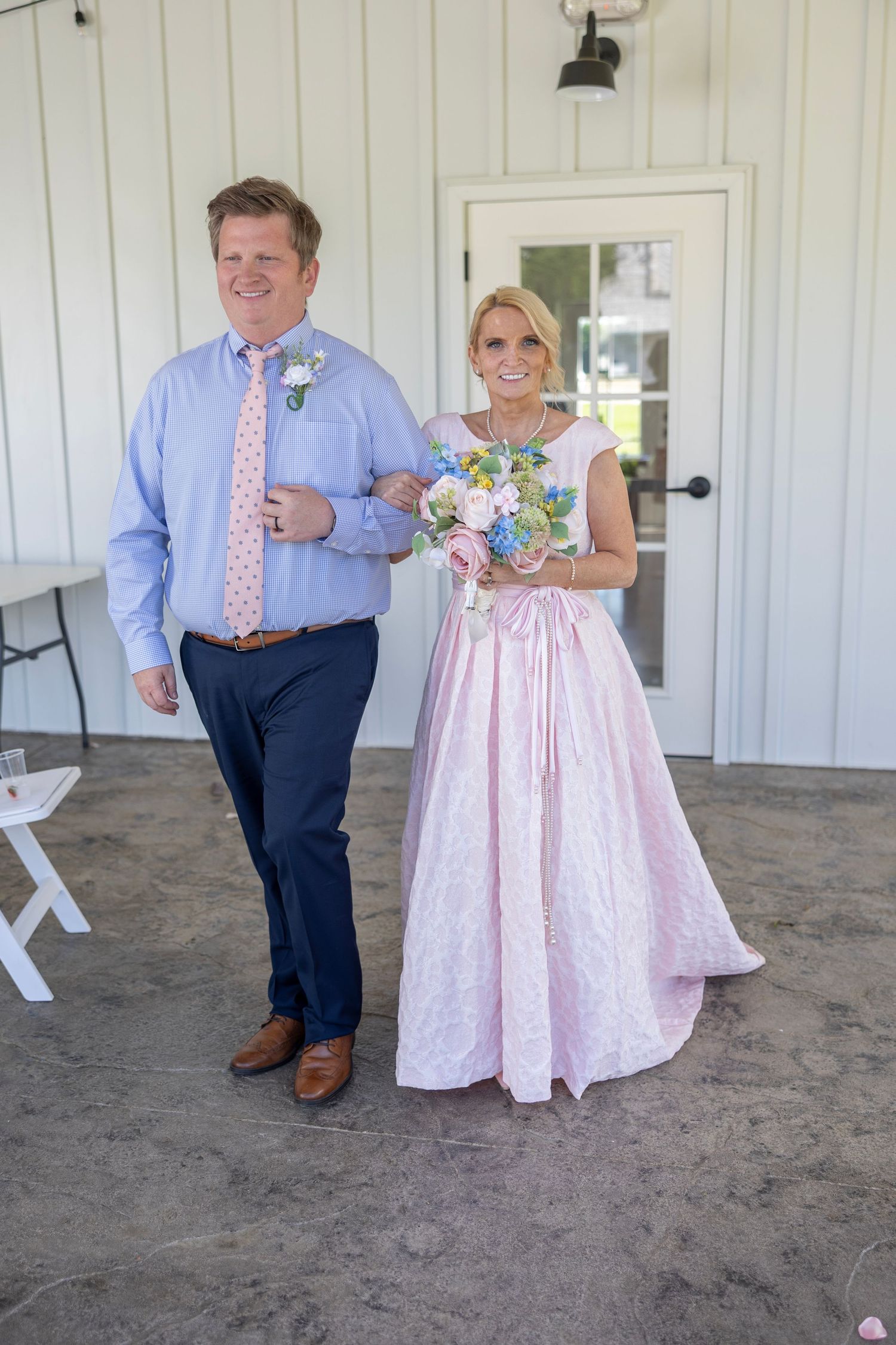 Couple in wedding attire, woman in pink gown holding bouquet, man in blue shirt and navy pants, walking together outdoors.