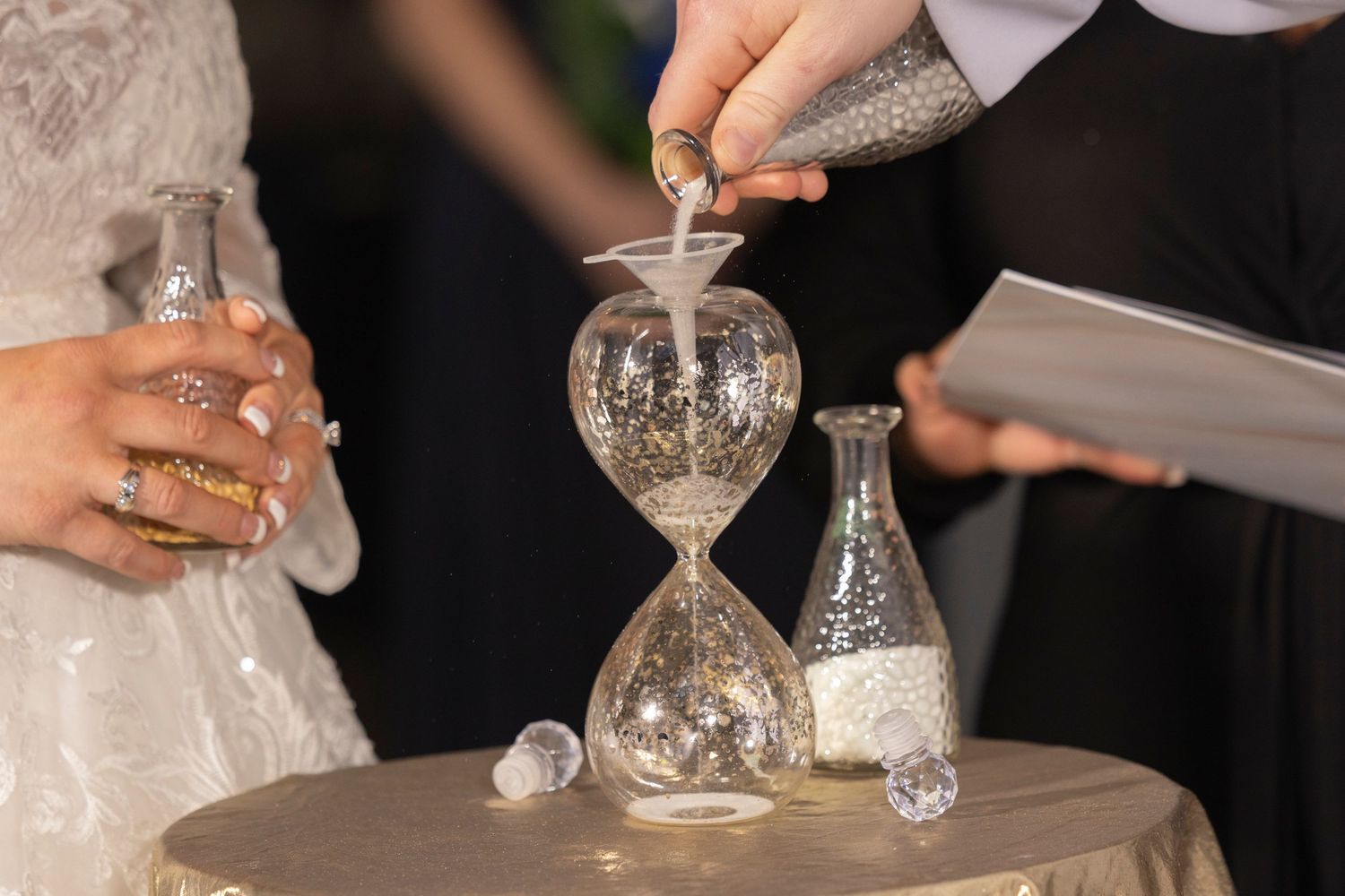 Wedding sand ceremony with couple pouring colored sand into hourglass vase on decorated table.