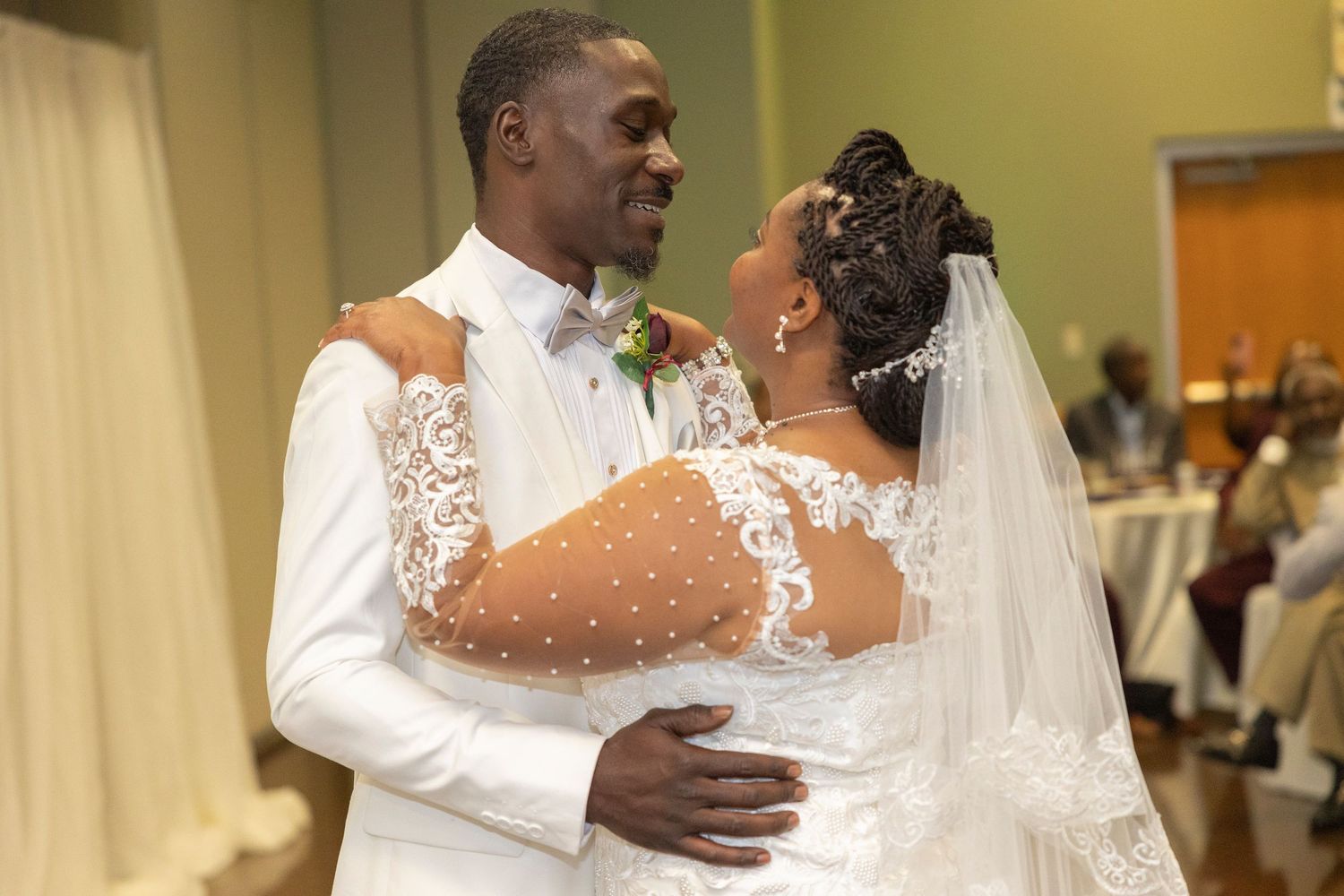 A bride and groom share their first dance, smiling at each other, bride in lace gown with veil, groom in white suit.