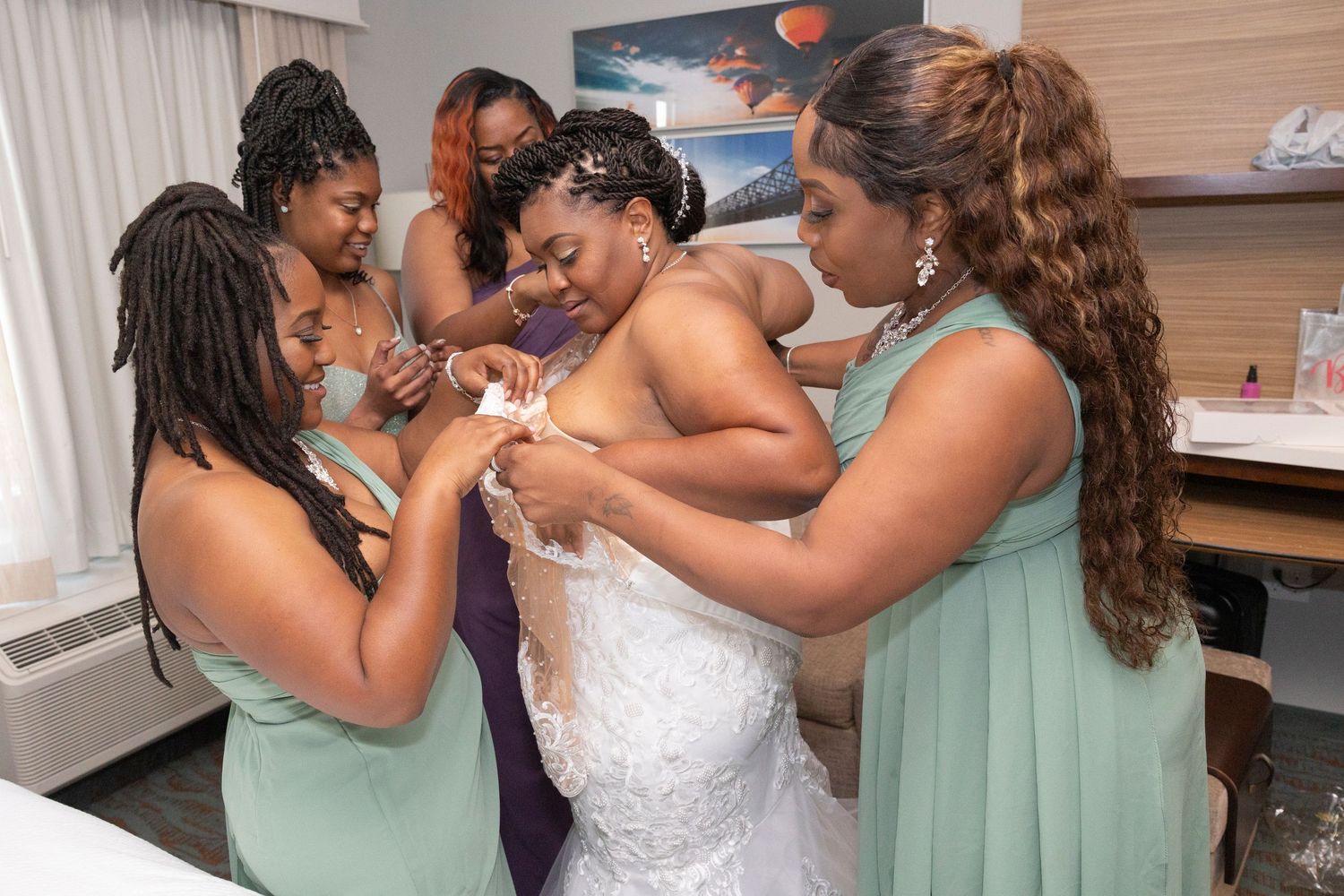 Bridesmaids in mint green dresses help a bride in a white lace gown get ready in a hotel room.