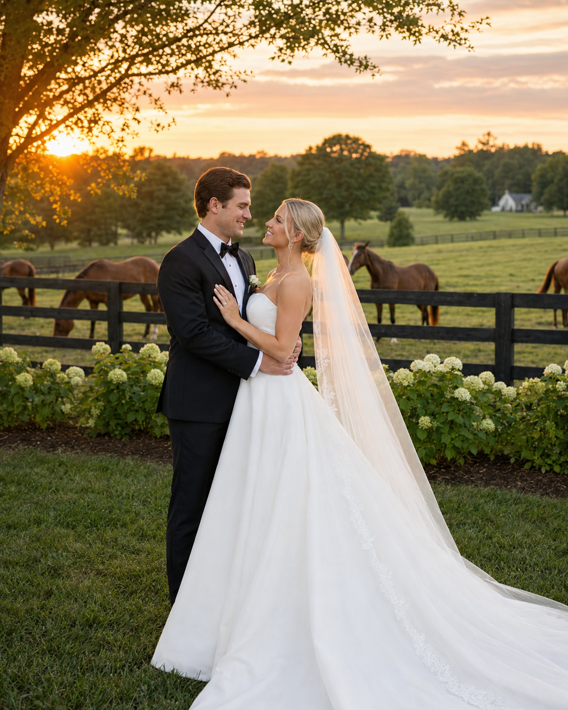 Bride and groom embrace at sunset on a lush farm estate with horses grazing behind a black fence.
