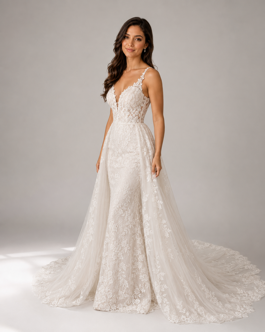 Elegant lace wedding gown with deep V-neckline, spaghetti straps, and detachable tulle overskirt train.