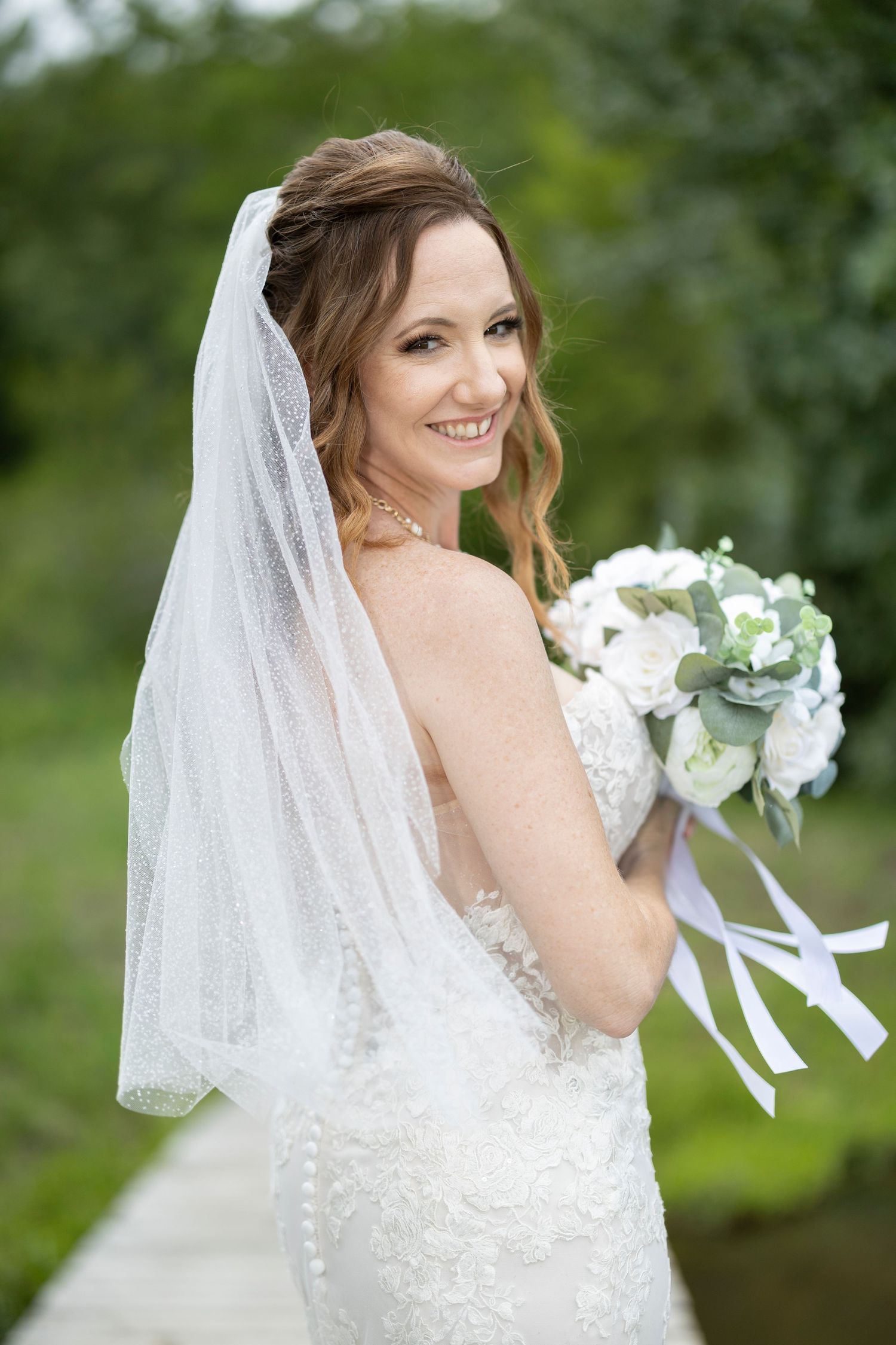 A bride in a white lace wedding dress and veil holds a white bouquet while posing outdoors against greenery.