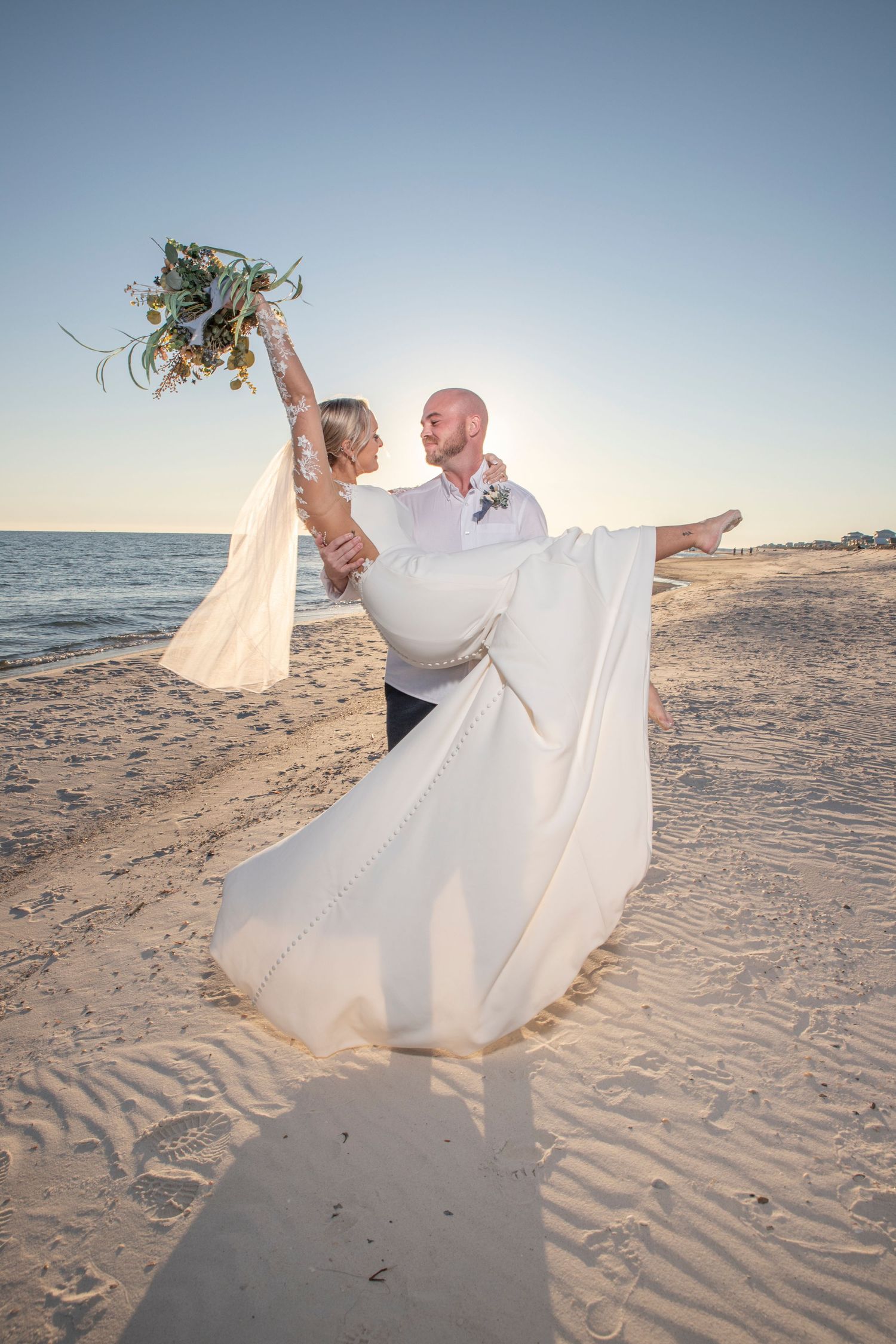 A couple in wedding attire dances together on a sandy beach at sunset, creating a romantic seaside moment.