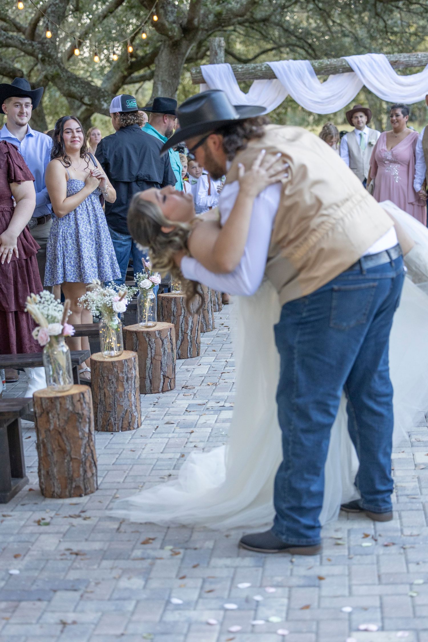 A couple performs a romantic dip dance move on a stone patio surrounded by rustic log pillars at an outdoor celebration.