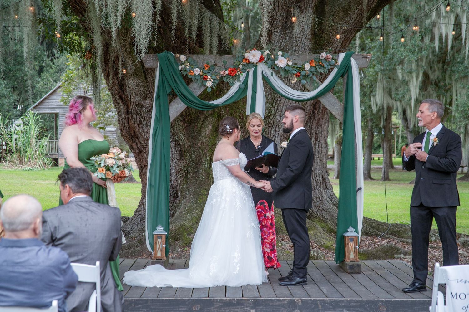 A wedding ceremony takes place under a rustic wooden gazebo surrounded by trees and hanging decorations.