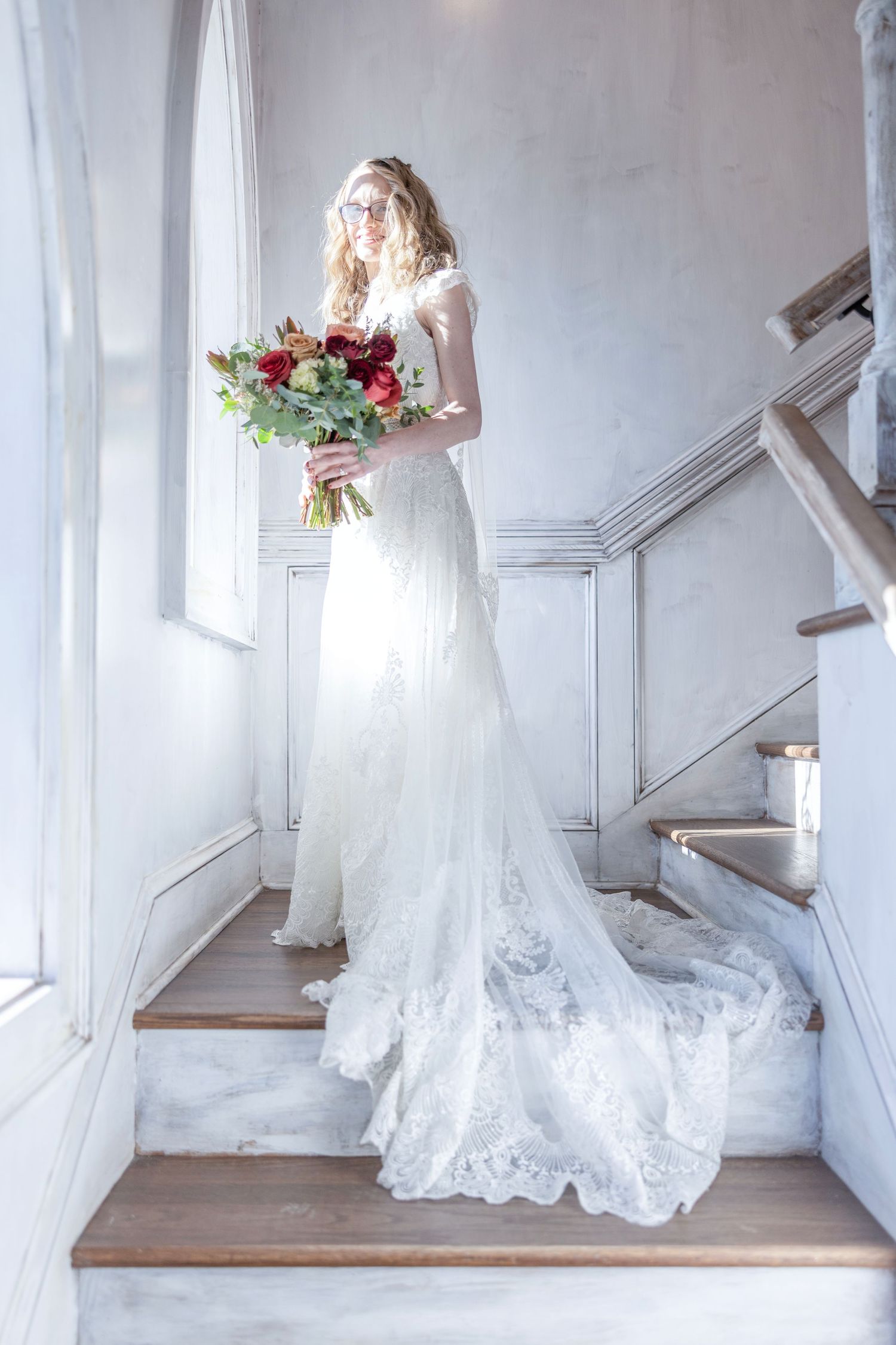 A bride in a white lace wedding dress with a veil stands in an elegant white hallway holding a red rose bouquet.