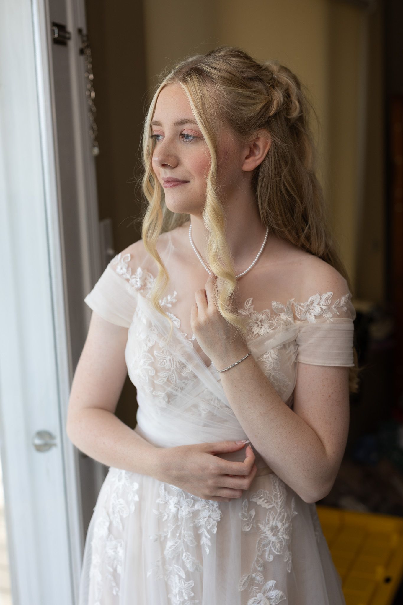 A bride in a delicate white lace wedding dress stands by a window in soft natural light.