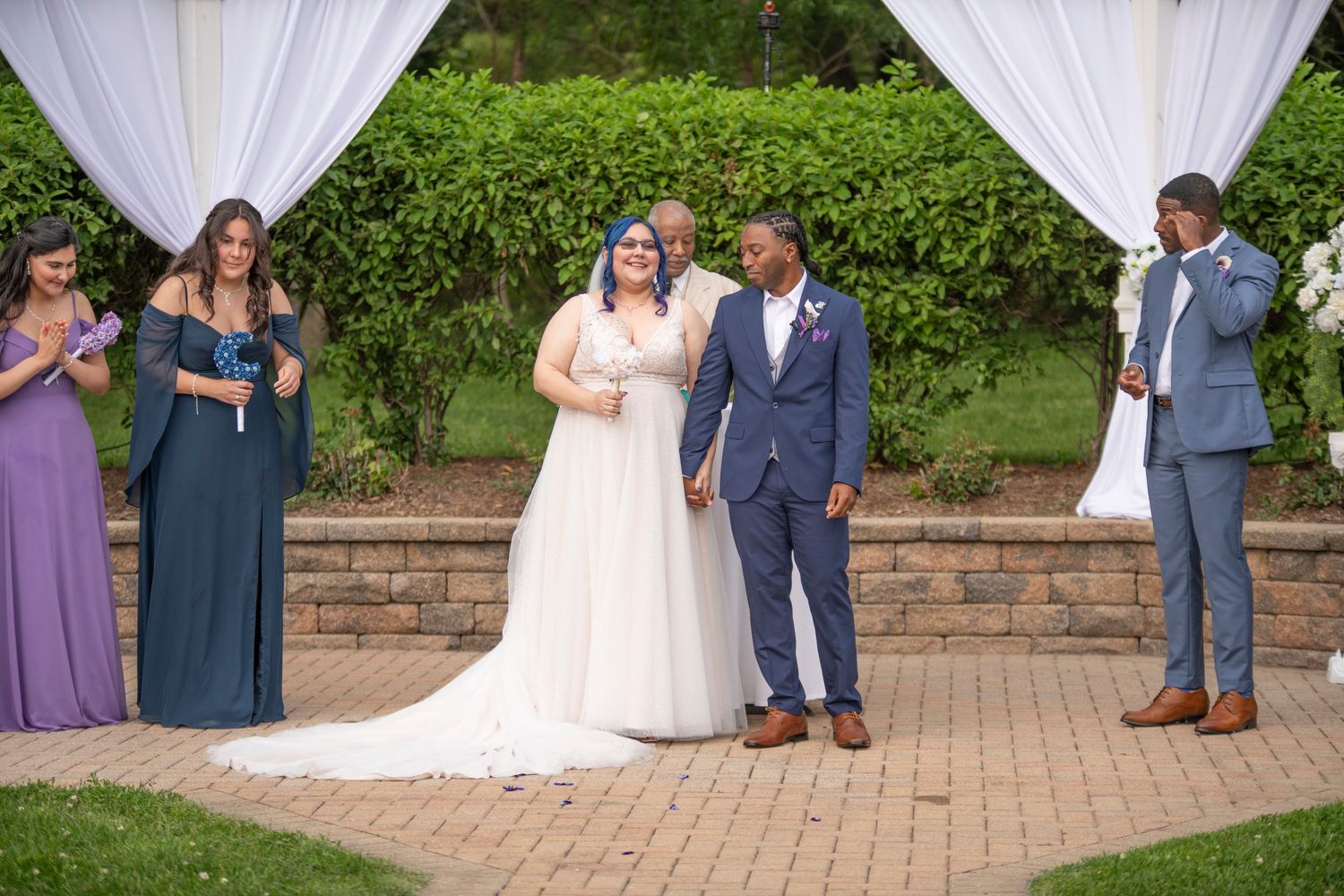 Wedding party stands together on brick patio surrounded by lush greenery in formal attire.
